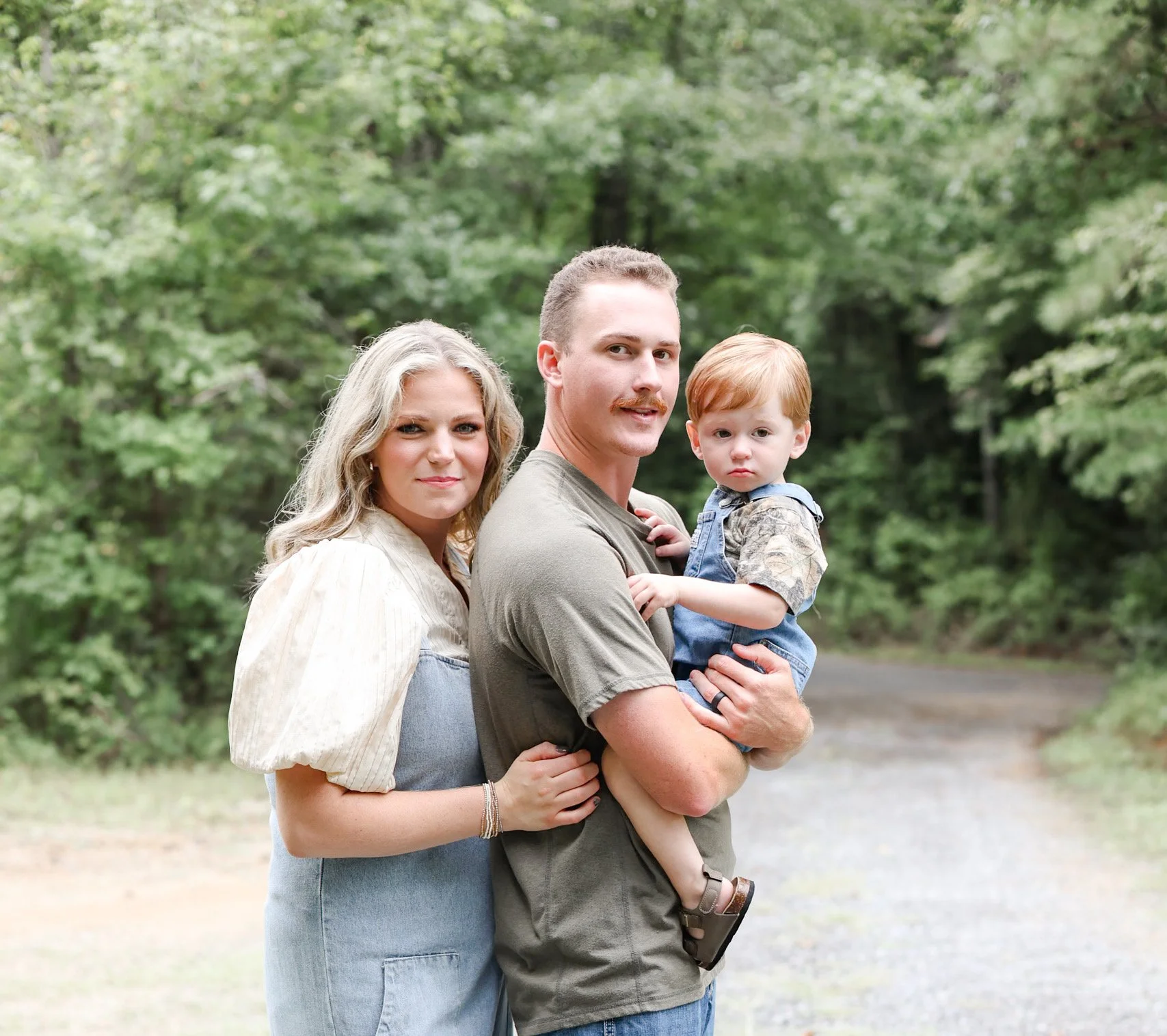 A family of three standing outdoors in front of green trees, with the father holding a young boy, and the mother standing close beside them.