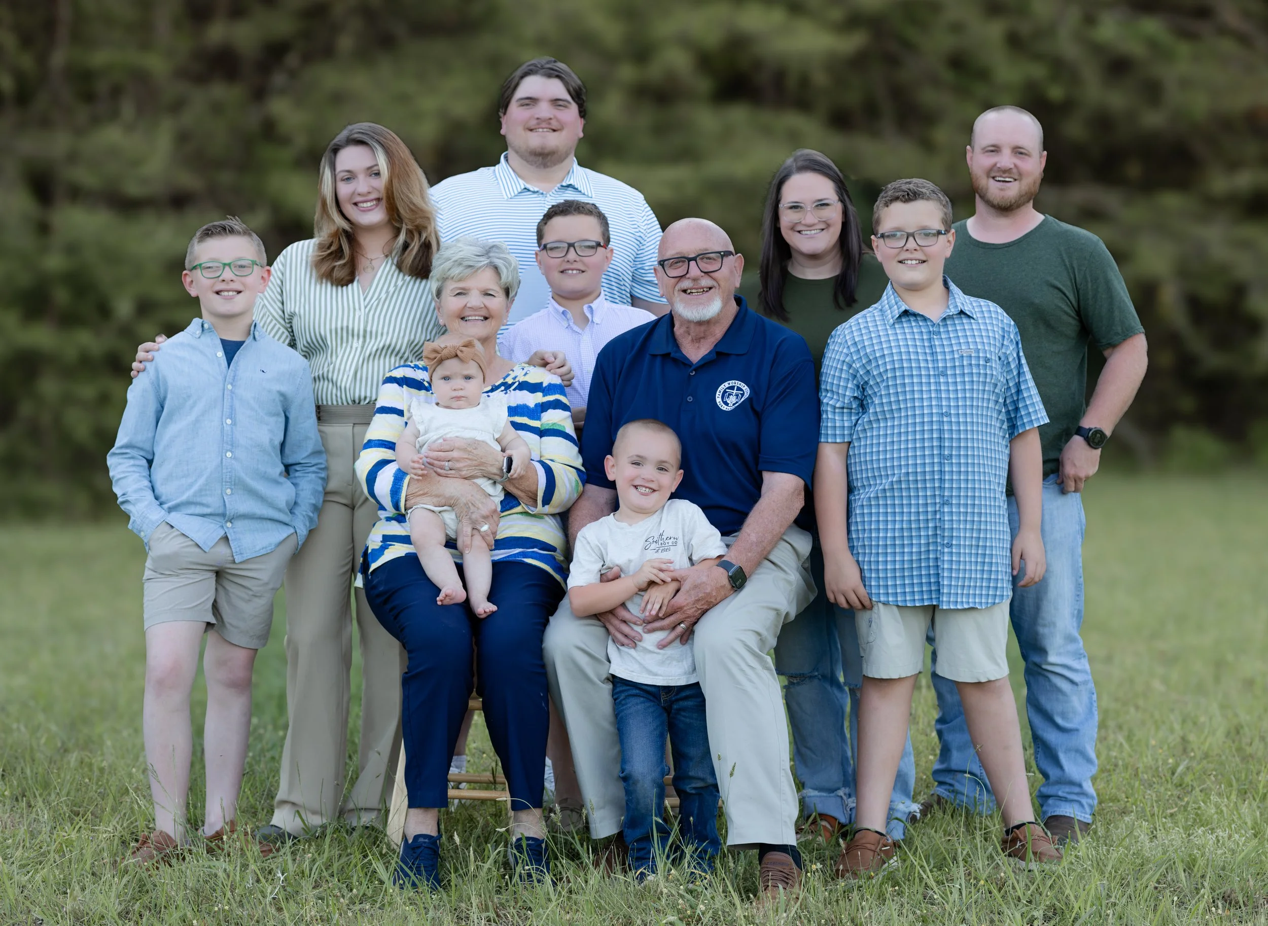 A multigenerational family of eleven people posing outdoors on a grassy area with a blurred green wooded background. They include children, teenagers, adults, and elderly individuals, all smiling and dressed casually.