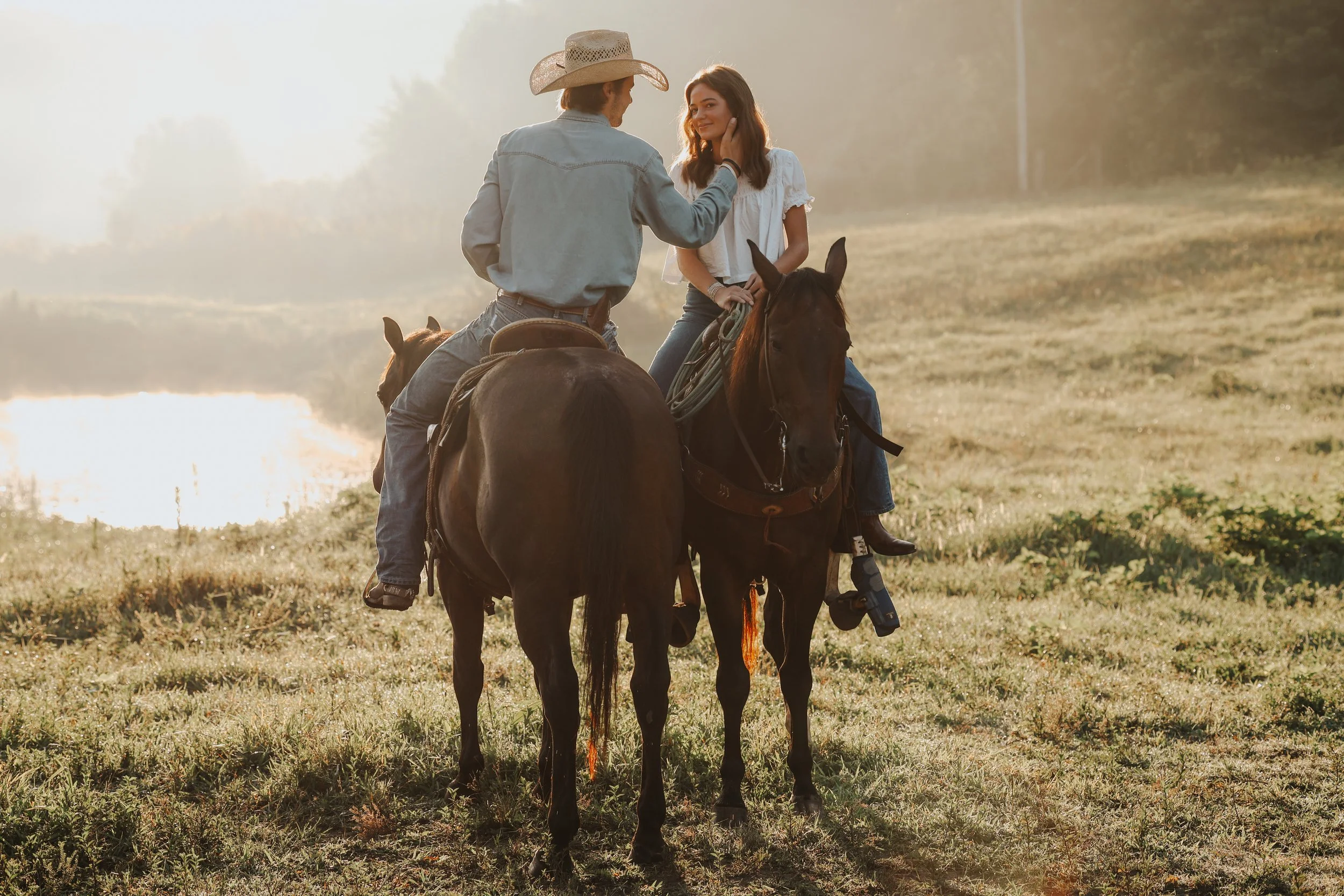 A man wearing a cowboy hat and denim shirt is riding a horse, touching a woman's face as she sits on another horse in an open field during sunset.