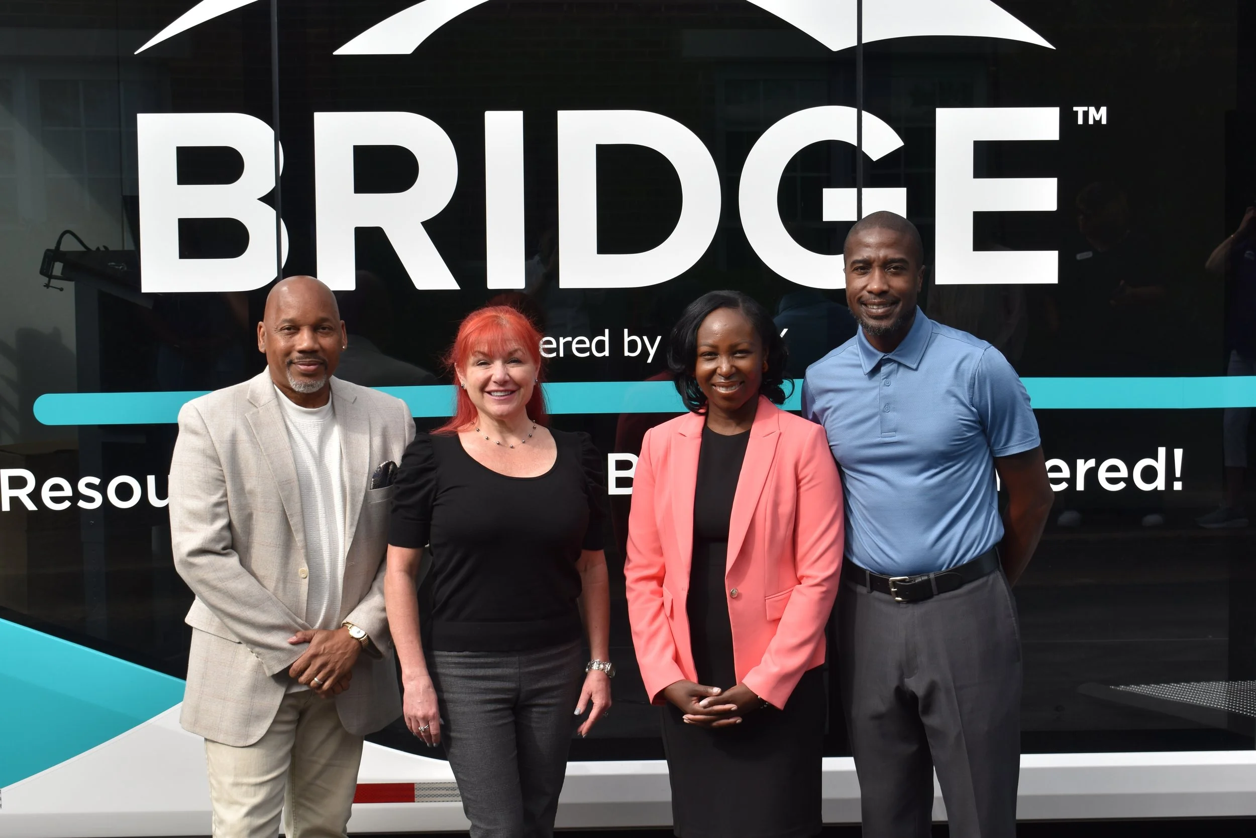Four diverse professionals standing in front of a large sign reading 'BRIDGE' during a public event. The group includes two men and two women dressed in business casual attire, smiling at the camera.