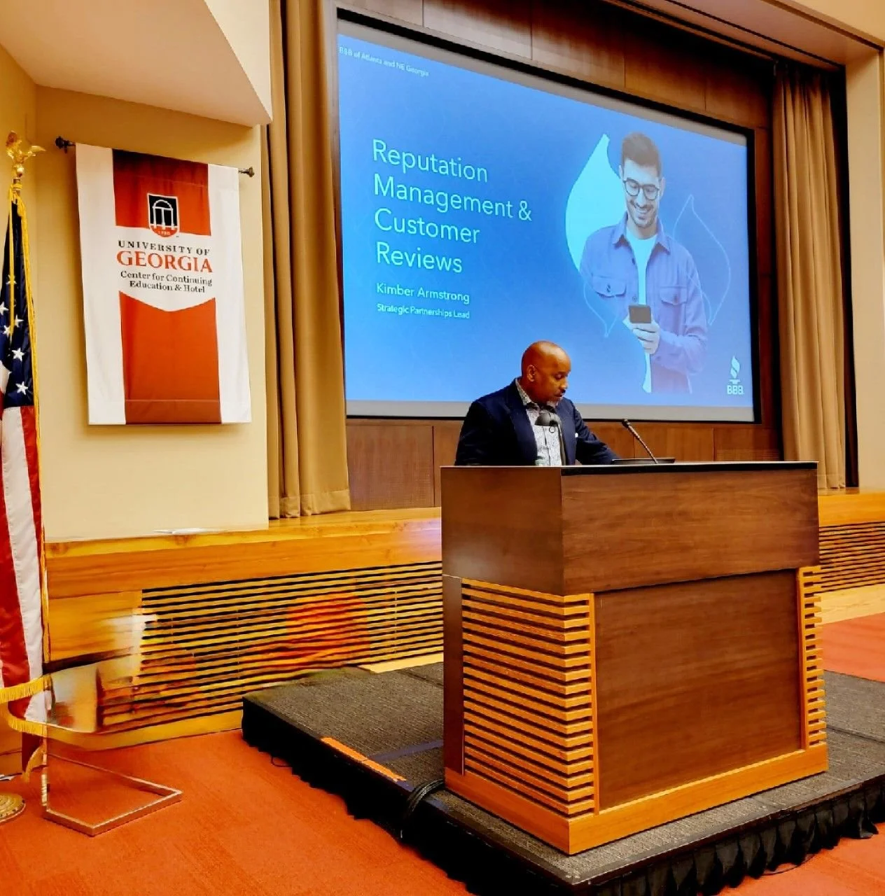 A man standing at a lectern giving a presentation on Reputation Management and Customer Reviews at the University of Georgia Center for Continuing Education & Hotel. A large screen behind him displays the presentation title. A banner with the University of Georgia logo is on the wall to the left of the screen.