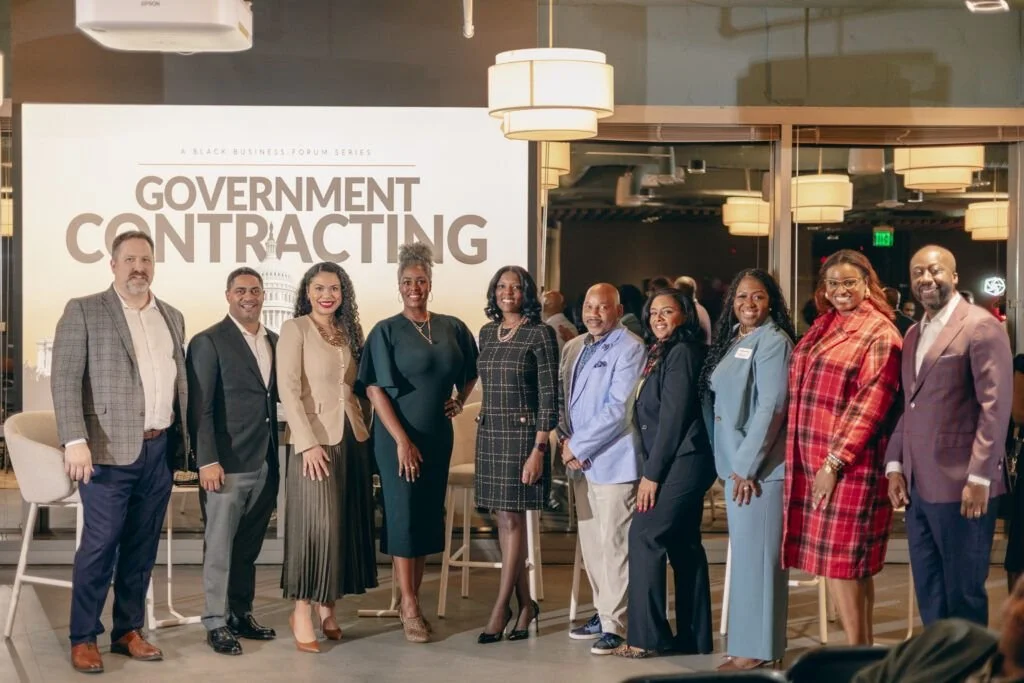 A group of ten diverse professionals standing together at an event titled 'Government Contracting,' with a large screen behind them displaying the event title. The setting appears to be a modern conference room with hanging lights.