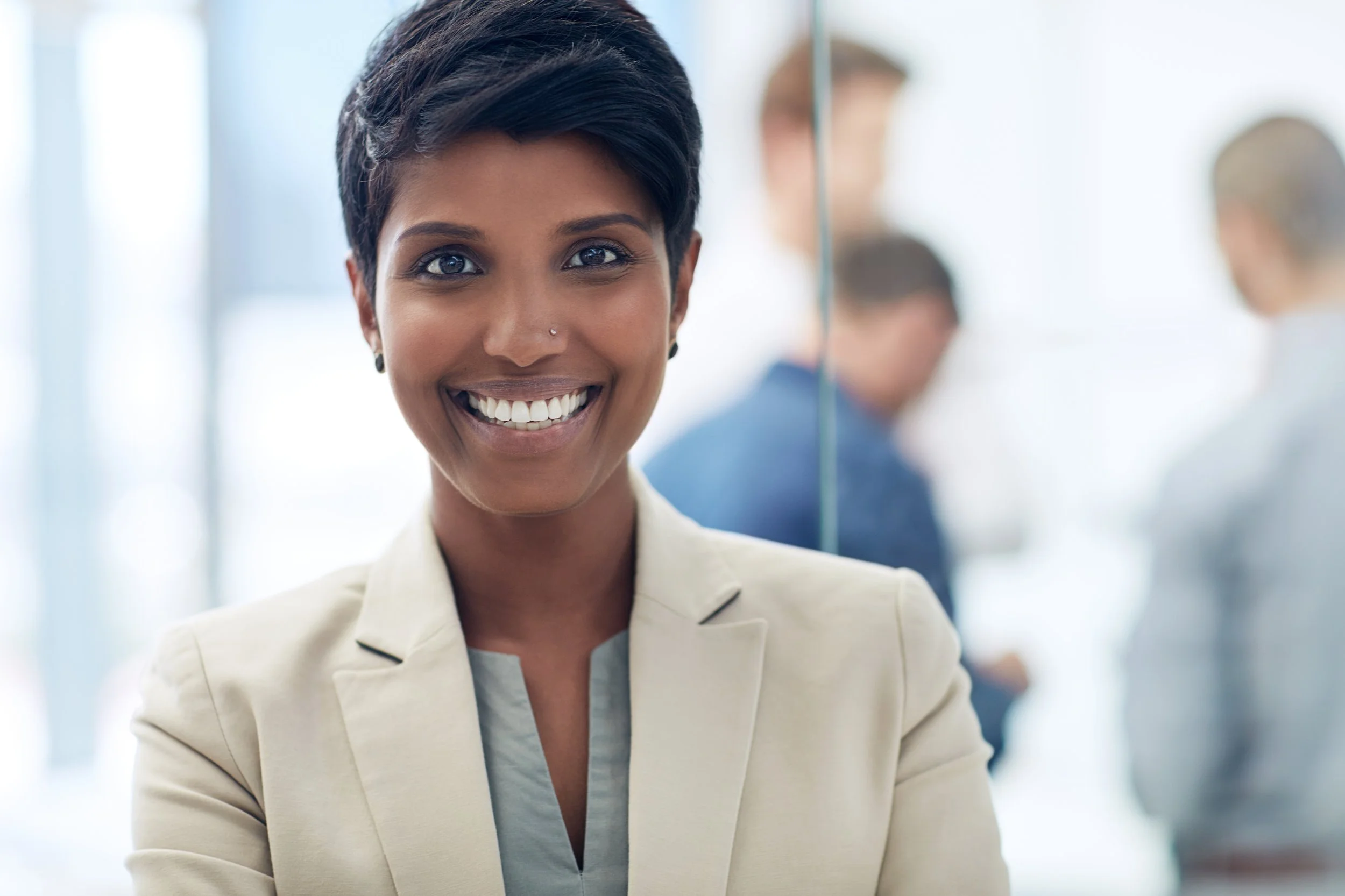 A woman smiling, dressed in a beige blazer, standing in a professional setting with other people blurred in the background.