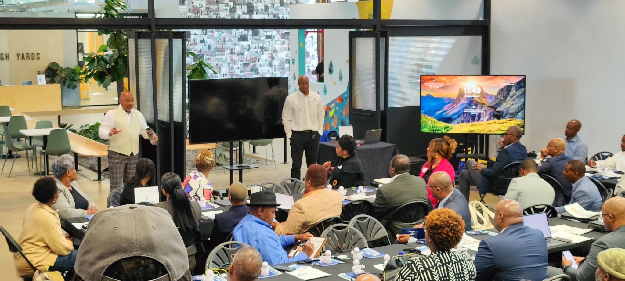 A diverse group of people attending a conference or workshop in a modern indoor space, with two men speaking at the front, large monitors displaying visuals, and attendees seated at tables with laptops and notebooks.