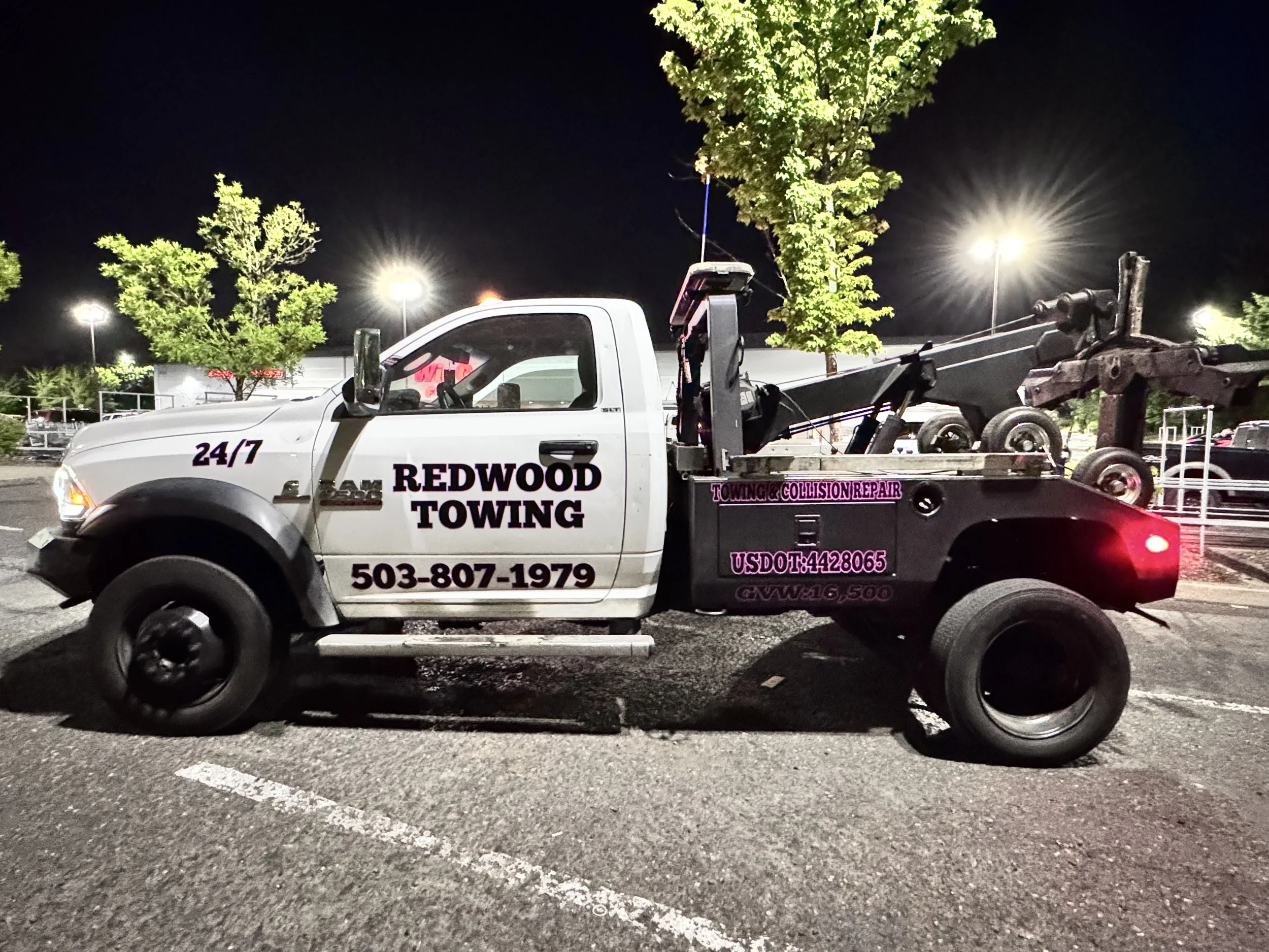 A white towing truck for Redwood Towing parked in a parking lot at night with trees and streetlights in the background.