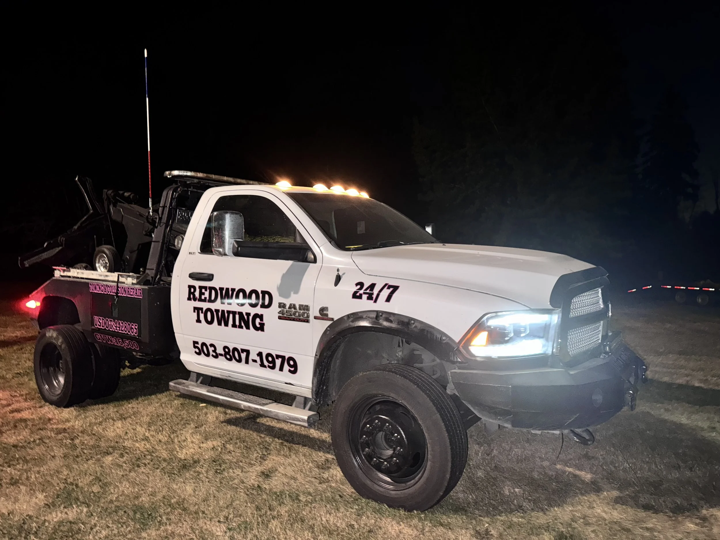 White towing truck with 'Redwood Towing' and contact information on side, front end damaged, at night with headlights on and tree silhouettes in background.