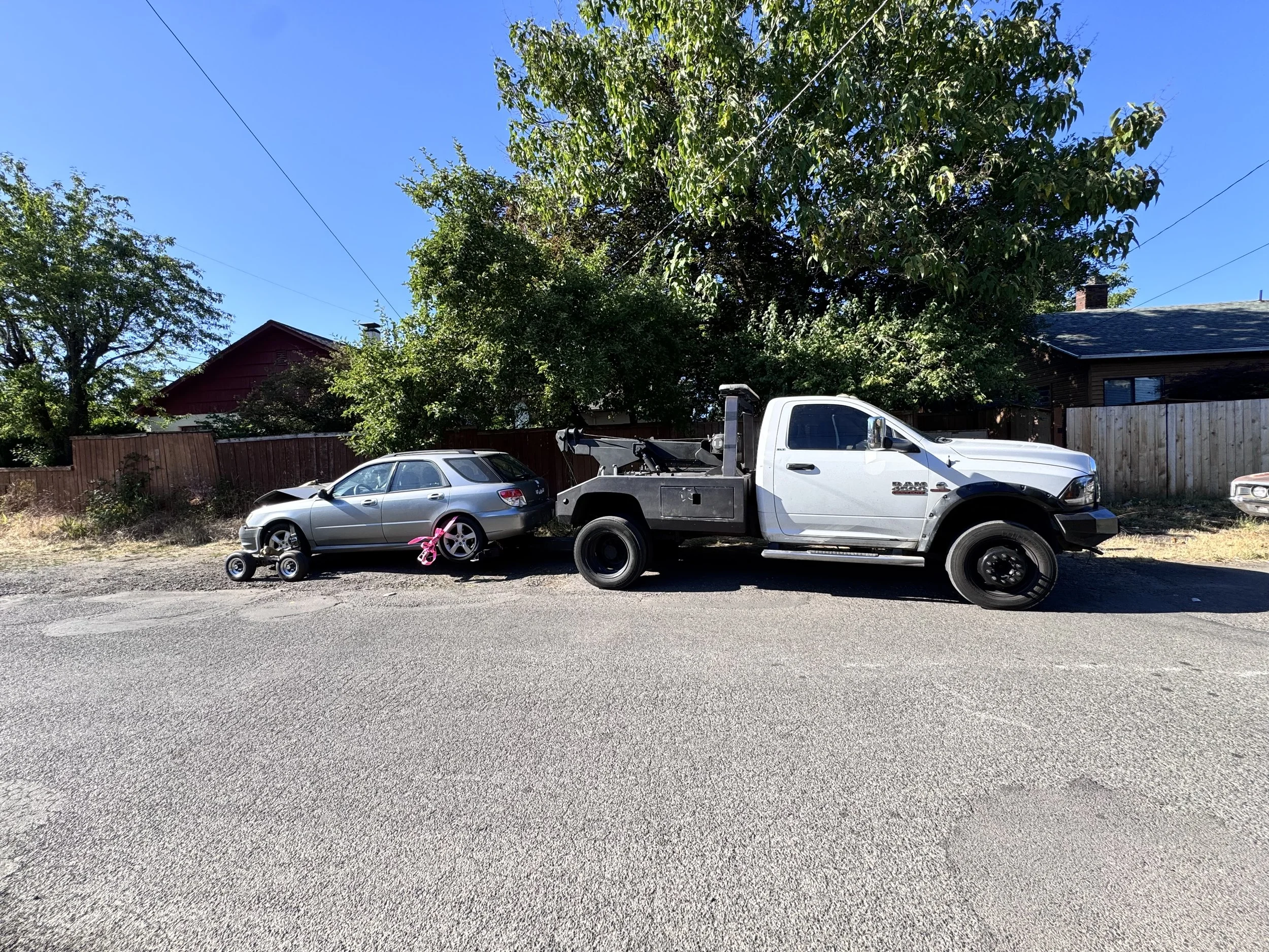 A silver sedan car and a white tow truck are parked on the side of a street. The sedan has a pink ribbon tied to its front. There is a small traffic cone near the front tire of the sedan. Fenced houses and green trees are in the background.