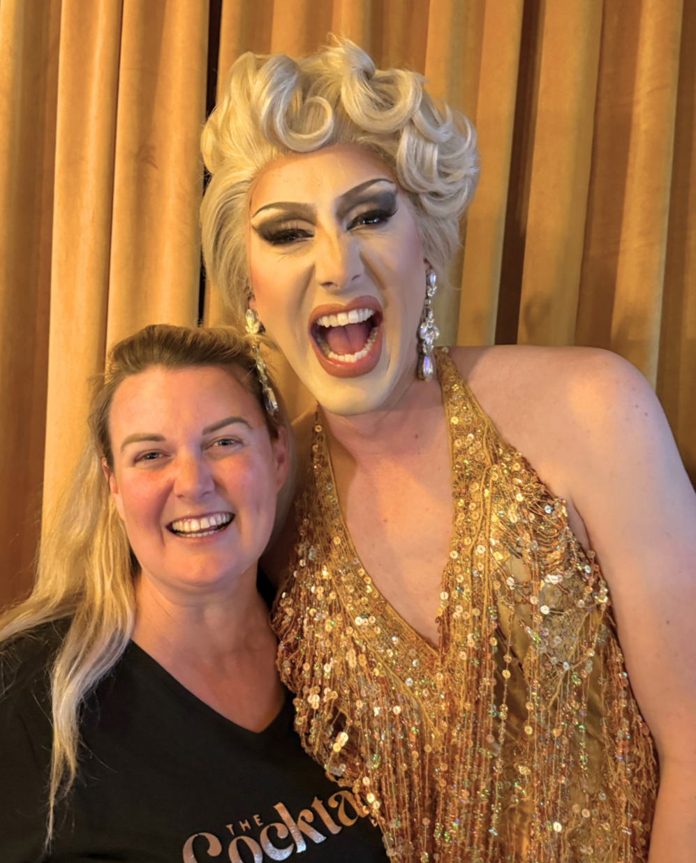Two women smiling and posing for a photo in front of a yellow curtain, with one dressed in a glamorous gold dress and the other in a black t-shirt.
