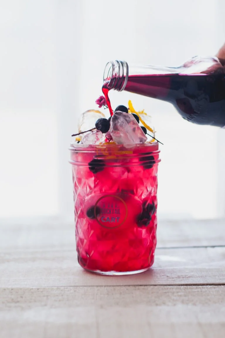 A glass jar filled with bright pink liquid, ice cubes, and black cherries, blackcurrant syrup being poured into it from a bottle, on a wooden surface.
