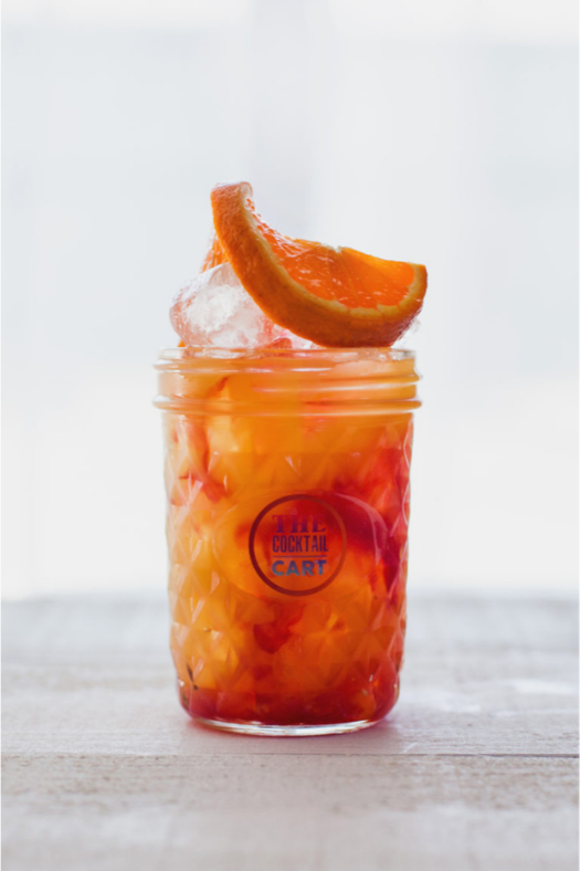 A glass jar filled with orange and red layered fruit cocktail, topped with an ice cube and a slice of orange