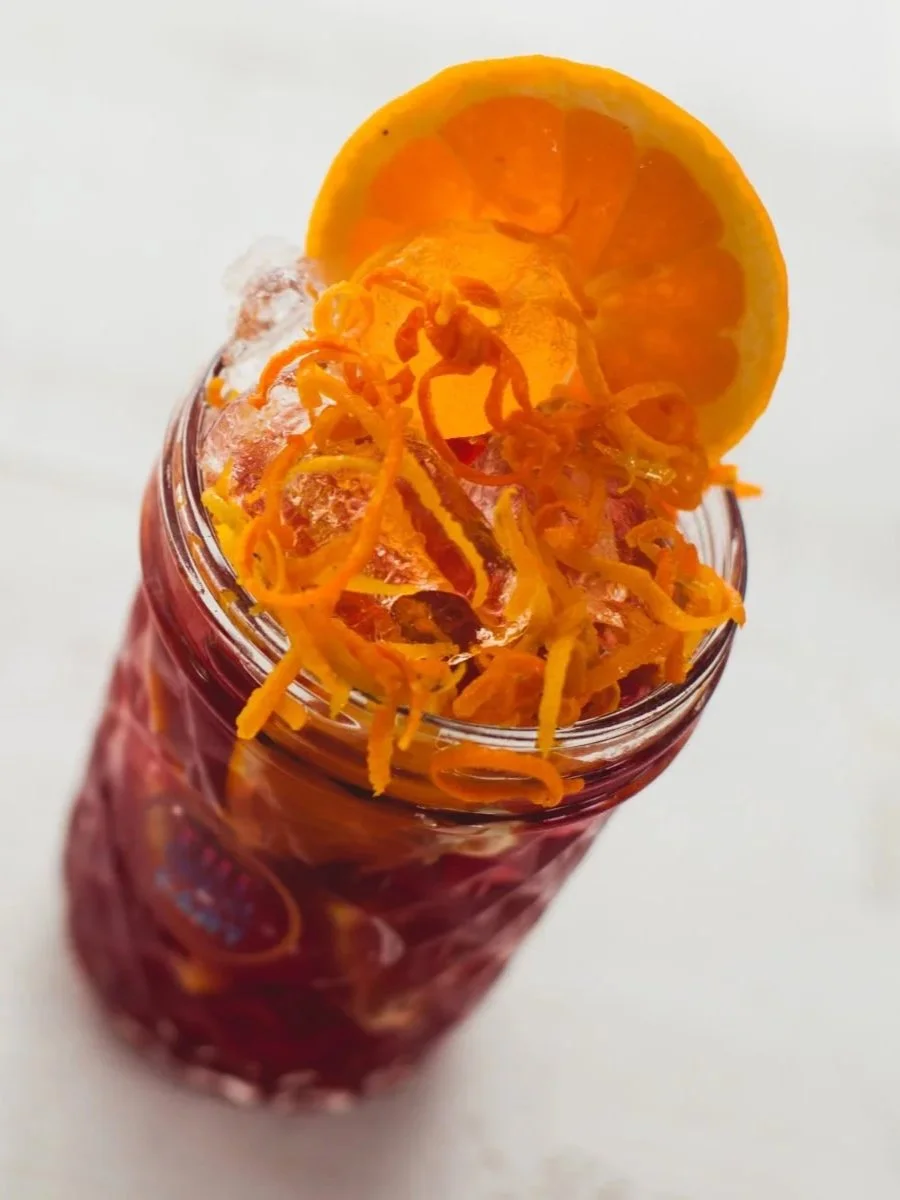 A glass jar filled with a red beverage, topped with shredded orange zest and a slice of orange.