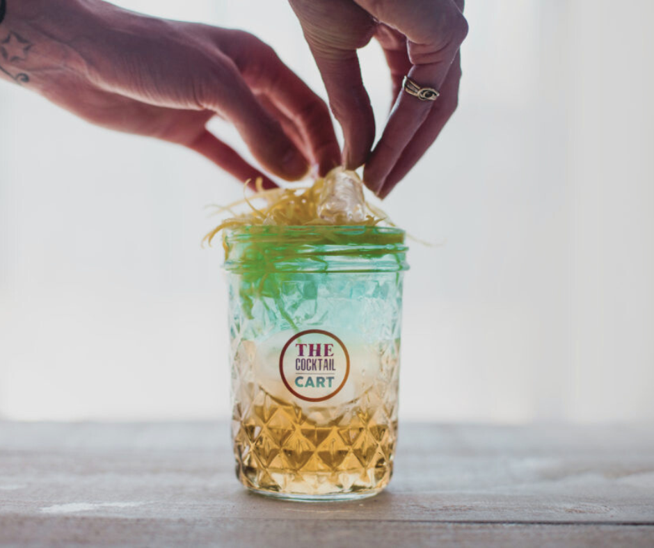 Hands placing a fresh lemon garnish into a decorative glass jar labeled "The Cocktail Cart" with a cocktail inside, on a wooden surface.