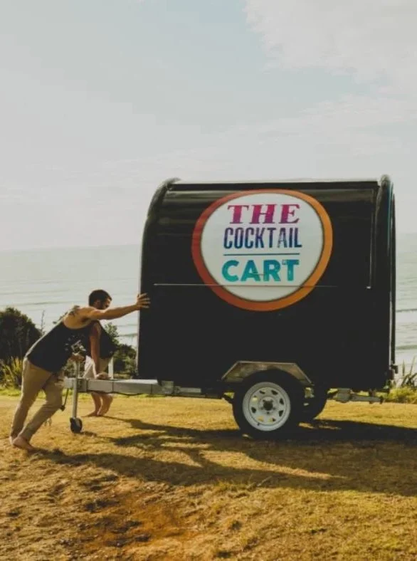 People pulling a small, black trailer with a circular sign that reads 'THE COCKTAIL CART' in colorful letters, outdoors on grassy terrain.