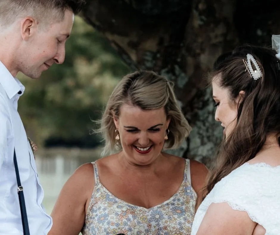 A woman is being a celebrant at a wedding between a man holding hands with a bride, who is dressed in a white gown, during an outdoor wedding ceremony.