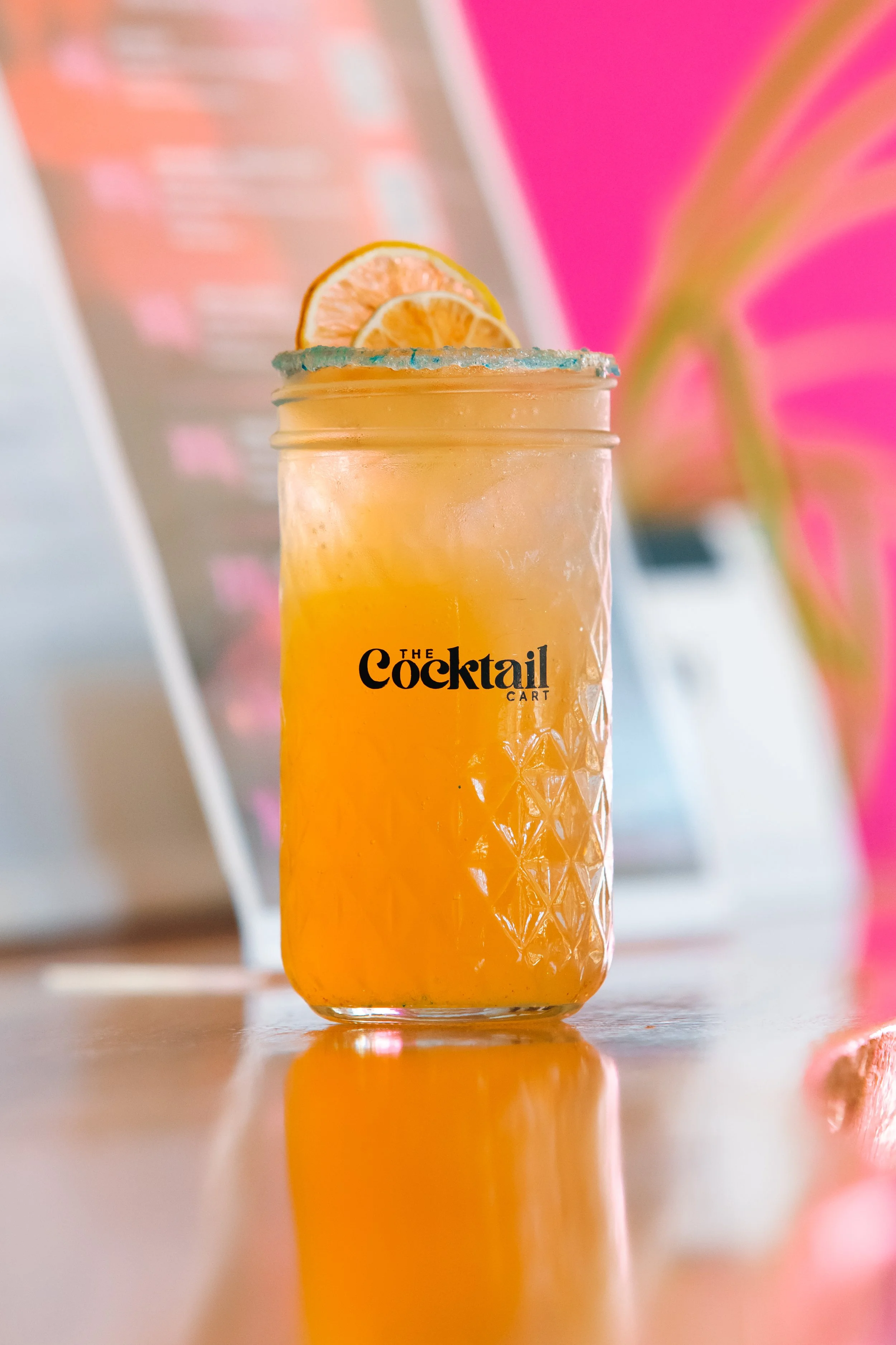 A glass jar with a blue sugar rim, filled with an orange-colored drink, garnished with two lemon slices, placed on a reflective surface with a pink and orange blurred background.