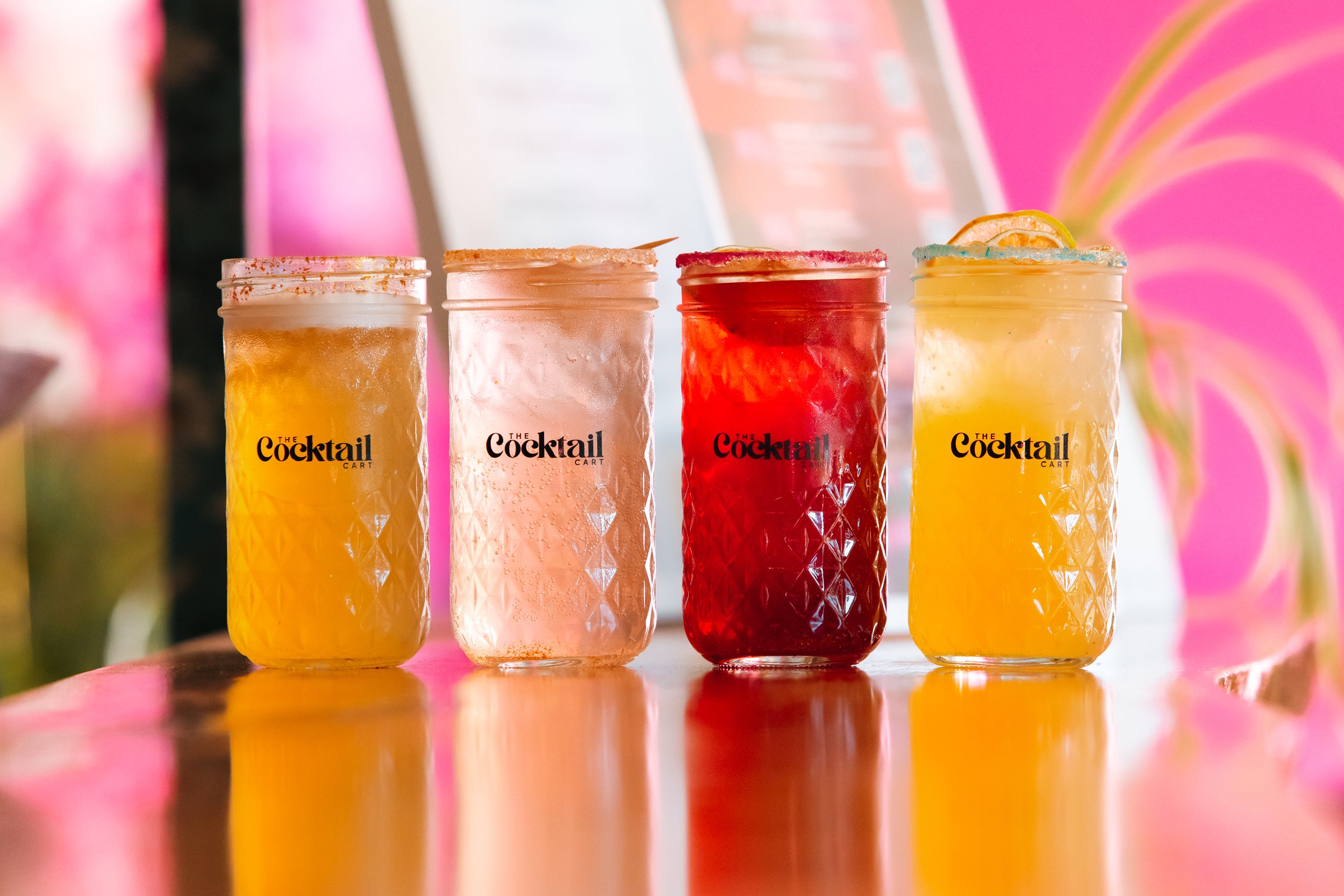 Four colorful cocktails in glass jars with textured surfaces, garnished differently, placed on a reflective surface with a pink and green background.