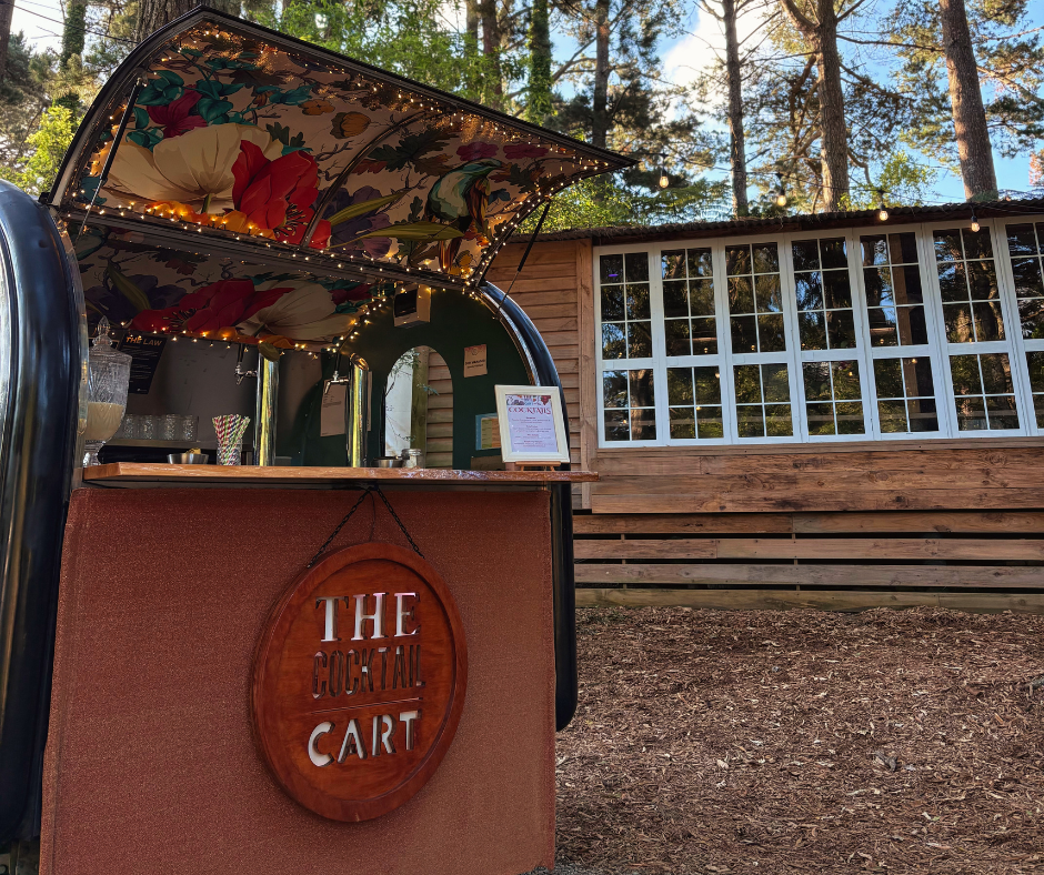 A cocktail cart decorated with string lights and a floral canopy, set outdoors near a wooden building with large windows, surrounded by trees.