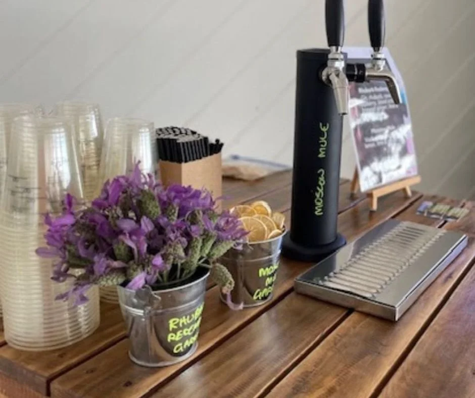 Mini cocktail bar with disposable cups, purple lavender flowers, a citrus slice bowl, a black tap with beverage, a metal scale, and a sign in the background.