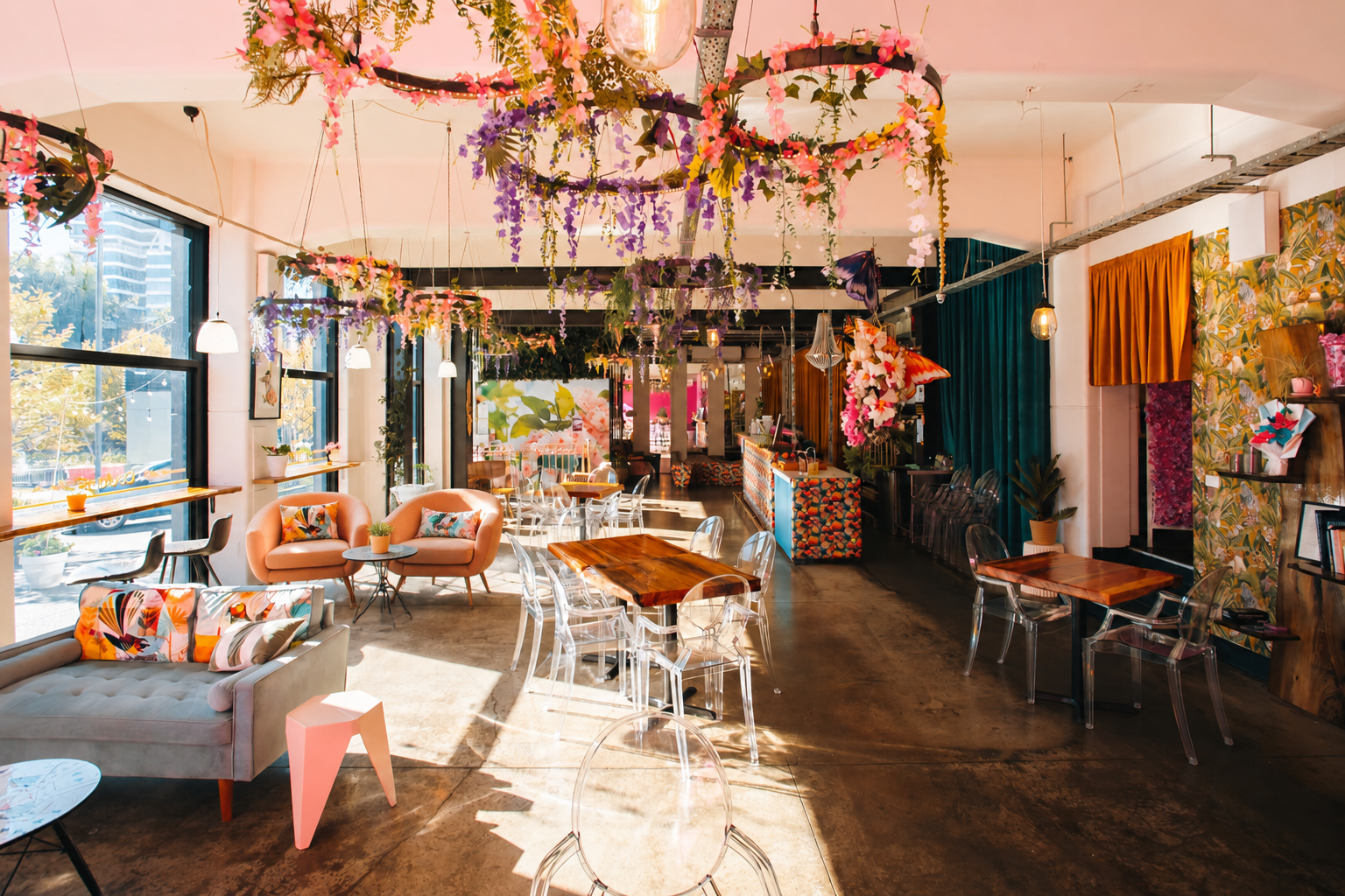 Colorful cafe interior with floral decorations, hanging pink and purple flowers, and sunlight streaming through large windows. It features pink and beige armchairs, clear acrylic chairs, wooden tables, and eclectic wall decor.