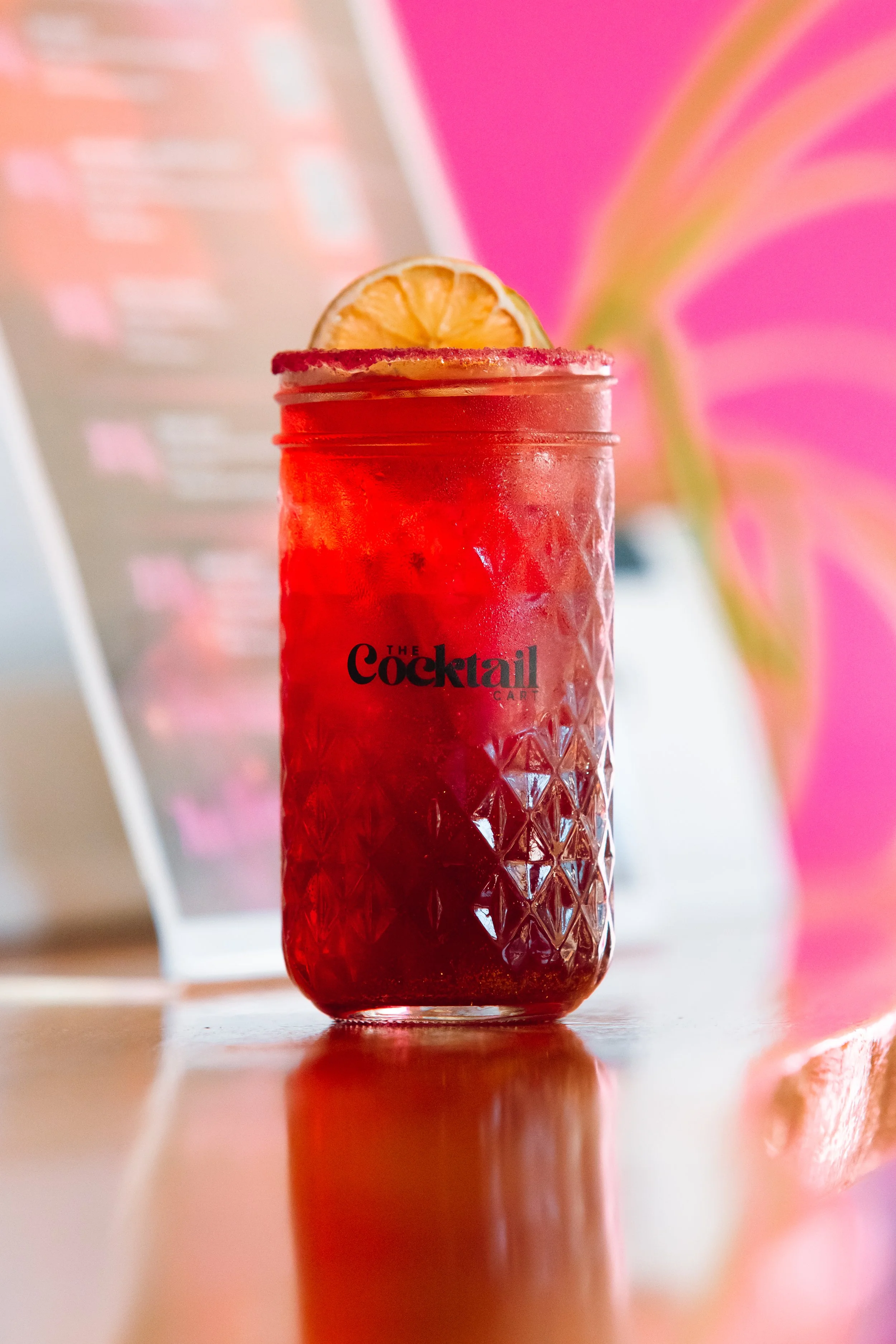 A red cocktail in a textured glass jar with a lemon slice garnish on the rim, placed on a wooden surface with a blurred menu or advertisement in the background.