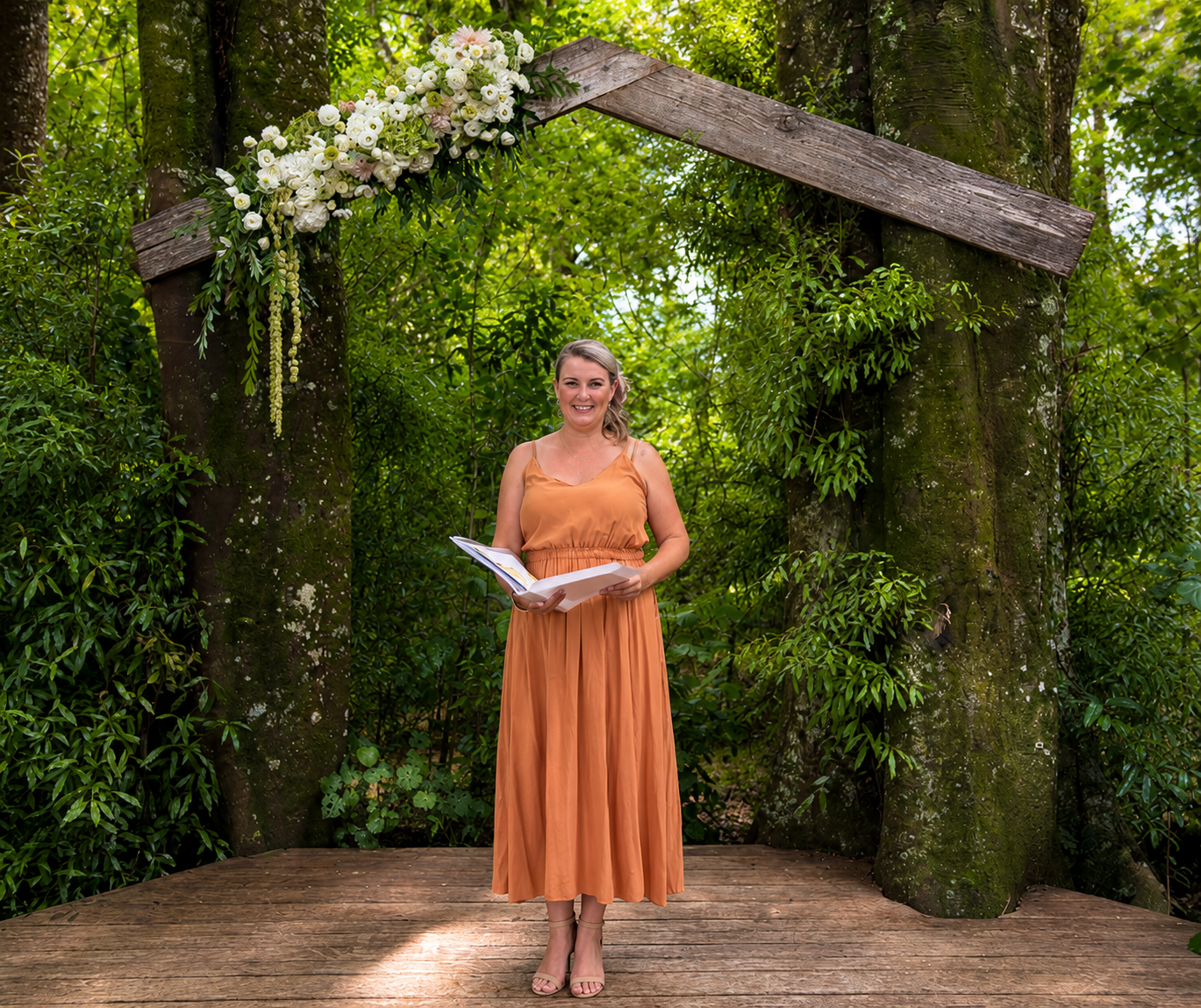 A woman in an orange dress holding a book, standing on a wooden platform under a decorated archway made of wood and adorned with white flowers and greenery, surrounded by trees and lush foliage in a forest setting.
