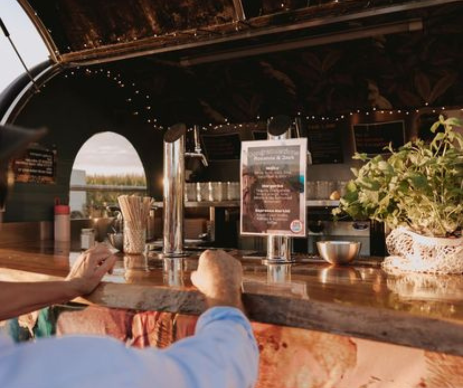 View of a bar counter with a person resting their arms on it, a menu sign, and potted plant, with a view of an outdoor landscape through an arched opening.