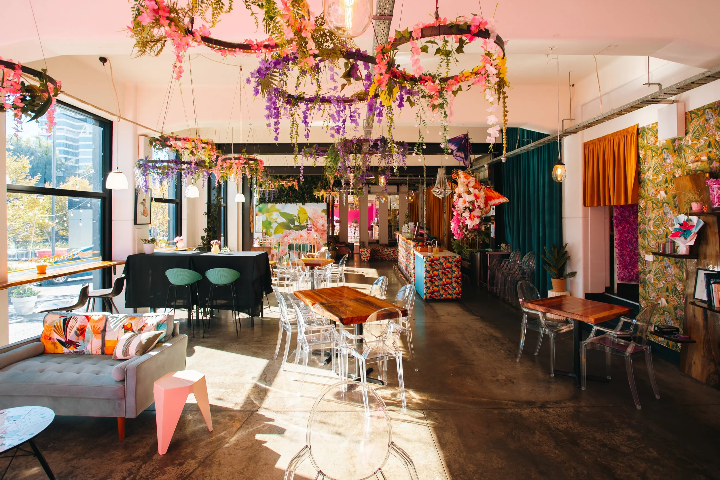 Interior of a bright, decorated café with hanging floral arrangements and colorful decor, including a floral sofa, various chairs, and large windows letting in natural light.