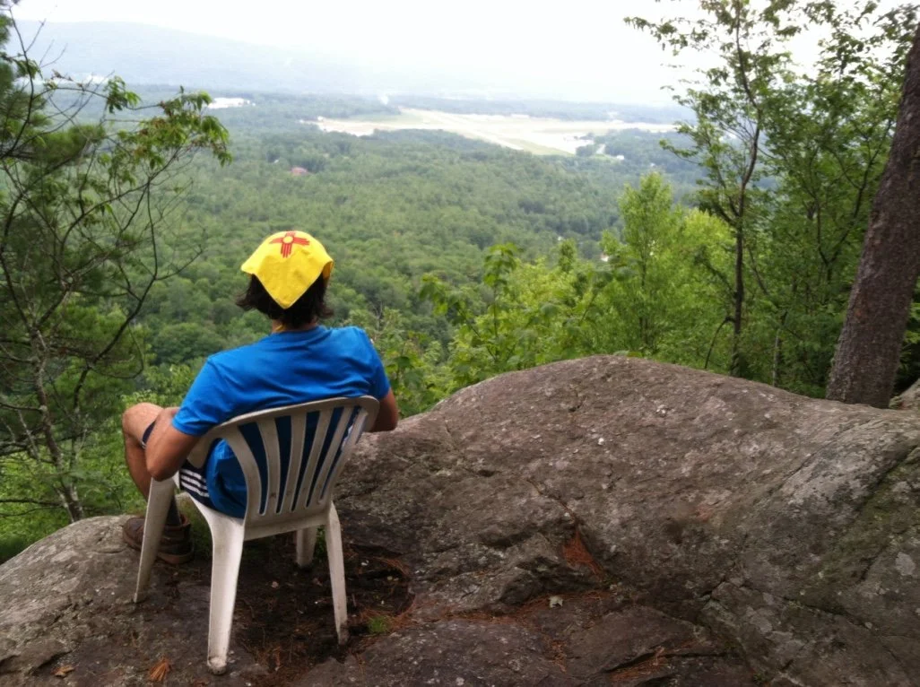 Man sitting on a white plastic chair on a large rock, overlooking a green forested valley and distant fields.