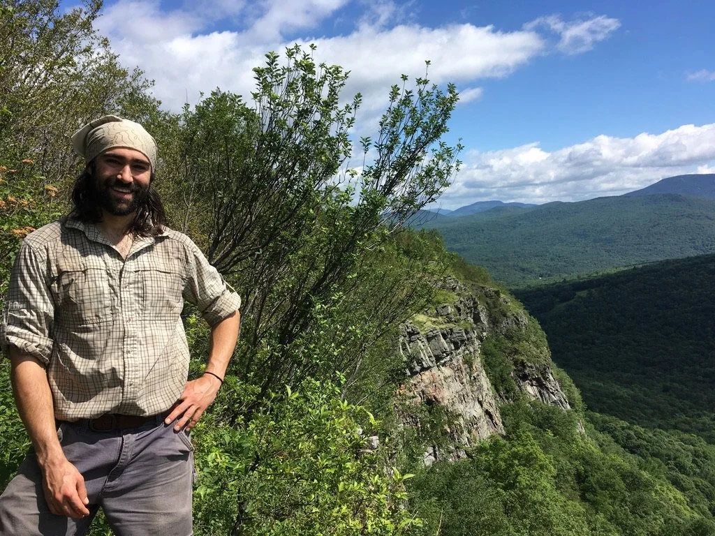 A man with long hair and a beard is standing outdoors on a hiking trail, wearing a beige plaid shirt, gray pants, and a beige headscarf, with a scenic mountain landscape and partly cloudy sky in the background.