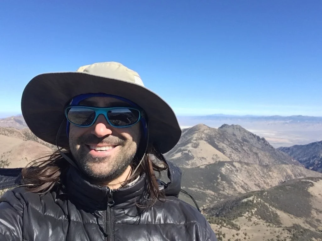 Smiling man with long hair, sunglasses, wide-brimmed hat, and black jacket taking a selfie with mountains and clear sky in the background.