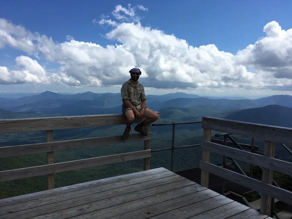 A man sitting on a wooden railing at a mountain overlook with a view of mountain ranges and a cloudy sky in the background.
