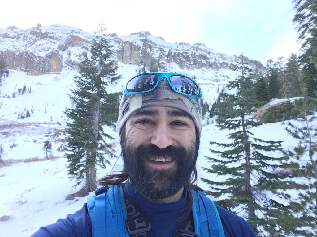Man taking a selfie in a snowy mountain landscape with pine trees and rugged cliffs in the background.