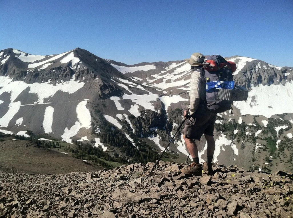 Hiker standing on rocky terrain overlooking snow-capped mountains under a clear blue sky.