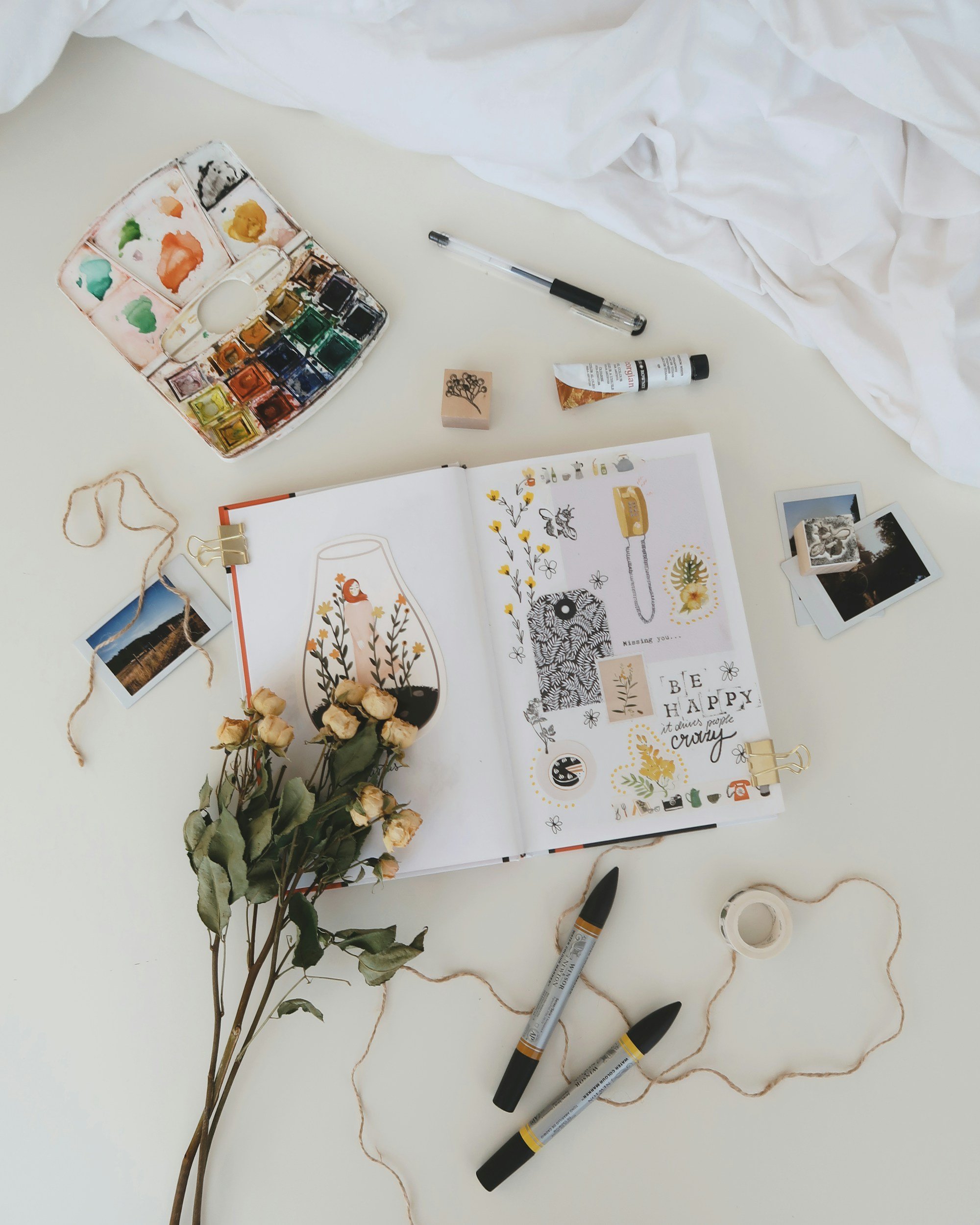 Open sketchbook with illustrations and decorative stickers, surrounded by art supplies including markers, watercolor palette, and tape, with dried roses and small Polaroid photos on a white surface. Representing art therapy.