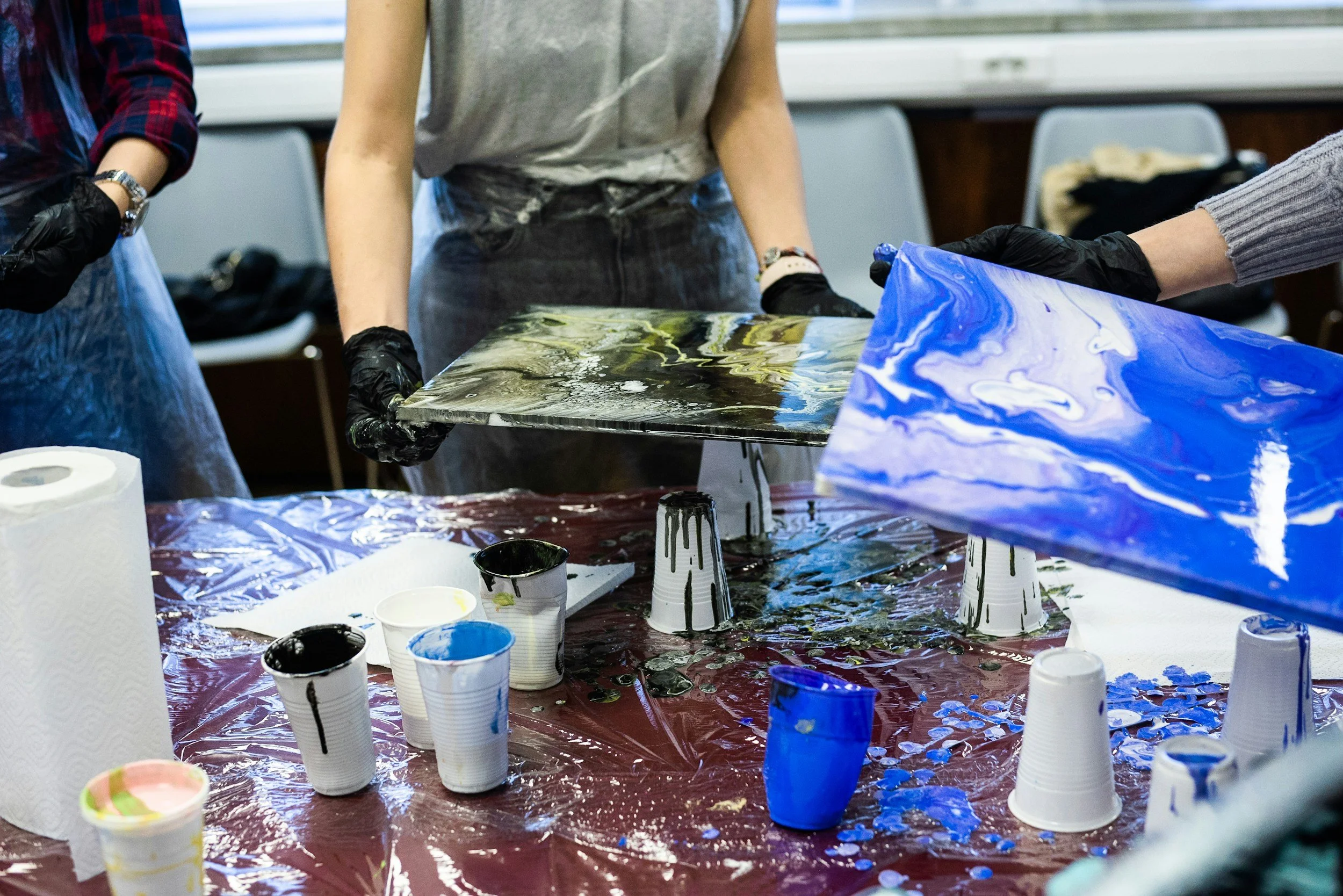 People creating abstract art with liquid paints on canvas using a marbling technique, with cups of paint and paper towels on a covered table.