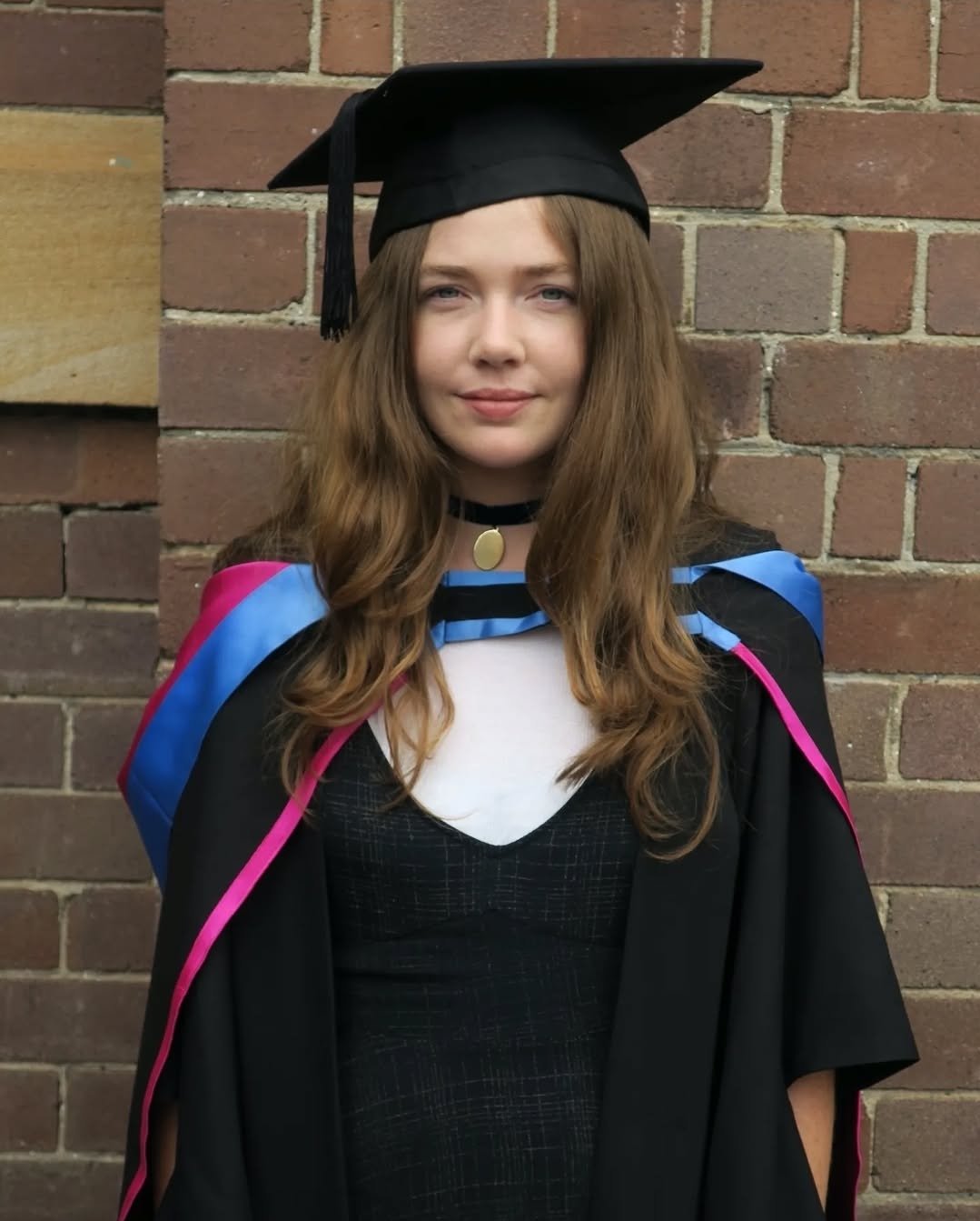 A young woman with long brown hair wearing a graduation cap and gown, standing in front of a brick wall.