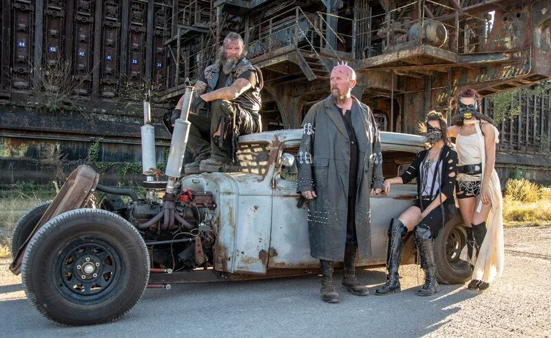 Group of five people in post-apocalyptic costumes standing next to a rusty, modified vehicle under a bridge.
