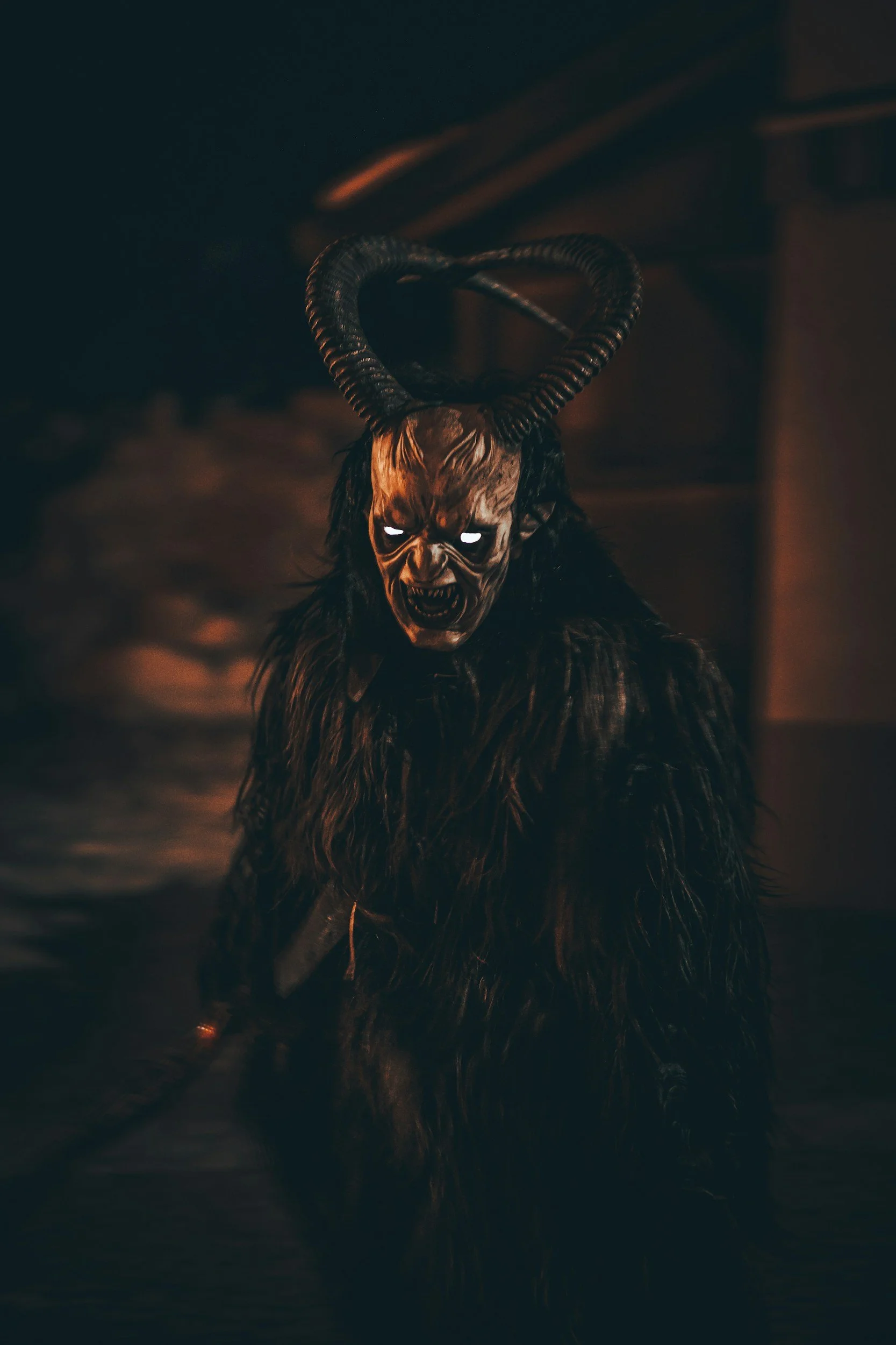 A frightening creature wearing a mask with glowing eyes, large horns, and a fur-covered body, standing in a dark, eerie setting.