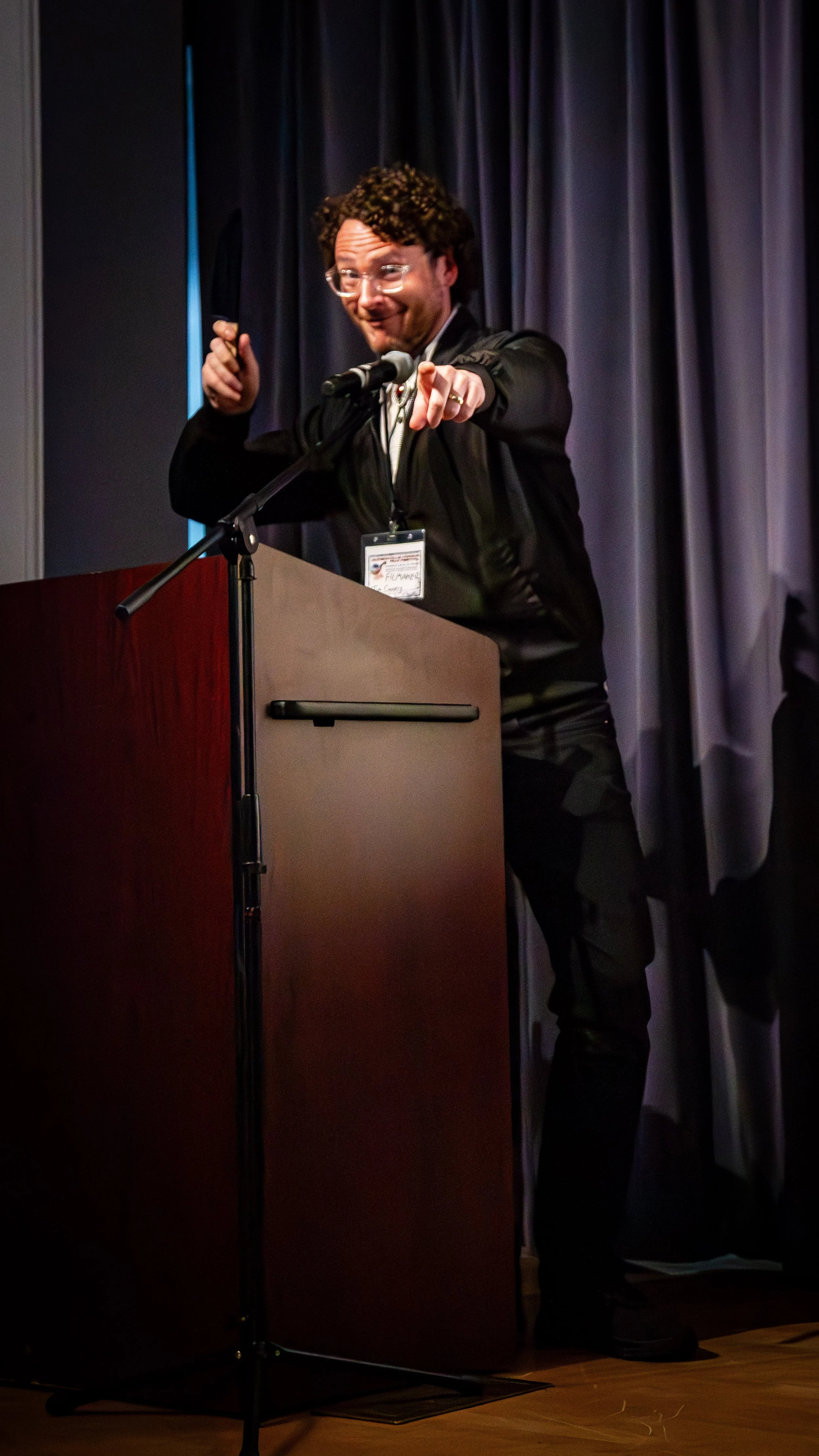 A man with curly hair and glasses standing at a podium, pointing forward and smiling, with a name badge hanging around his neck, in front of dark curtains.
