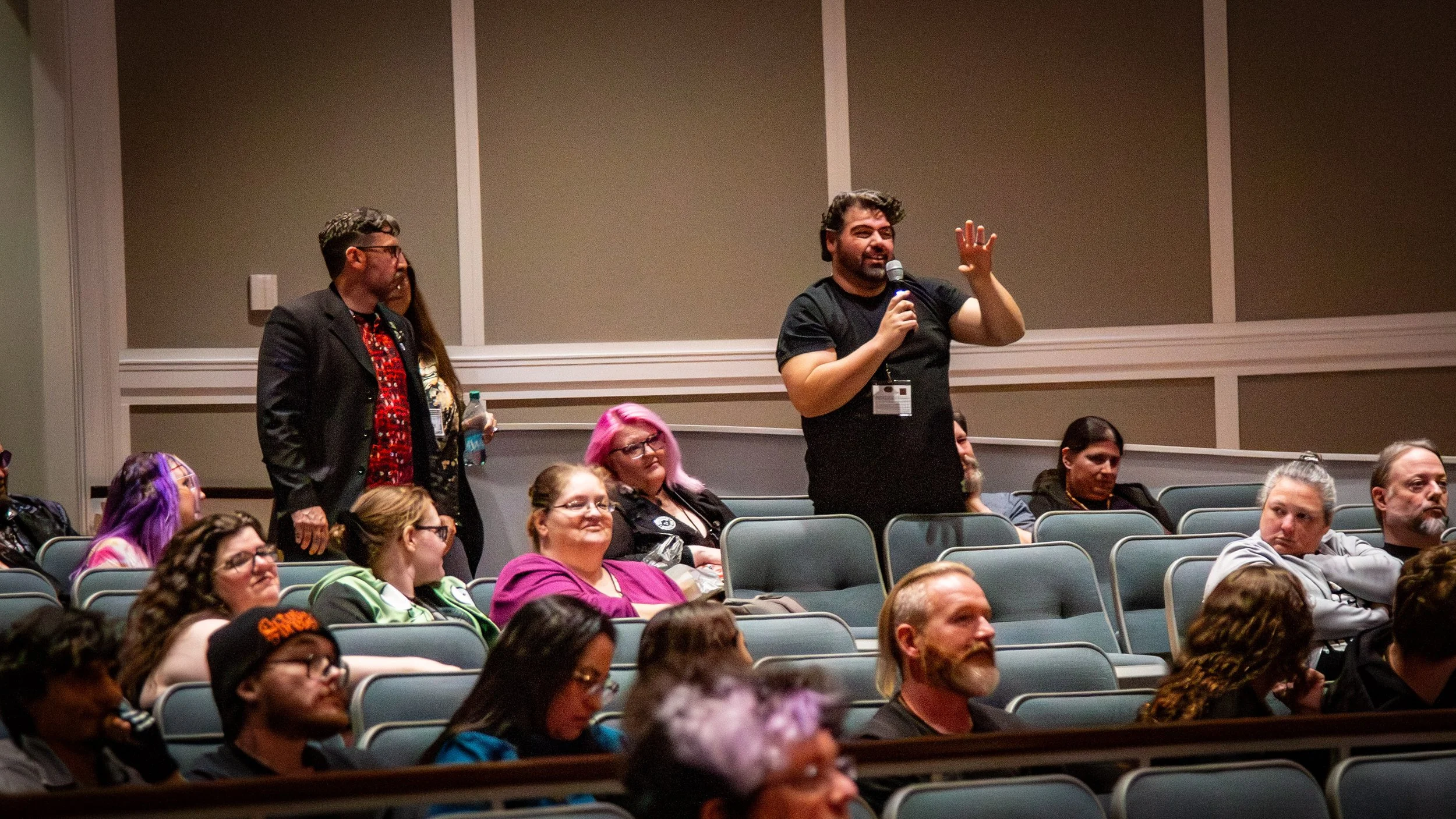 A man with dark hair and a beard stands up holding a microphone, speaking to an audience in a conference room. The audience is seated in rows, listening attentively, with some people appearing engaged and others more neutral. The room has beige walls