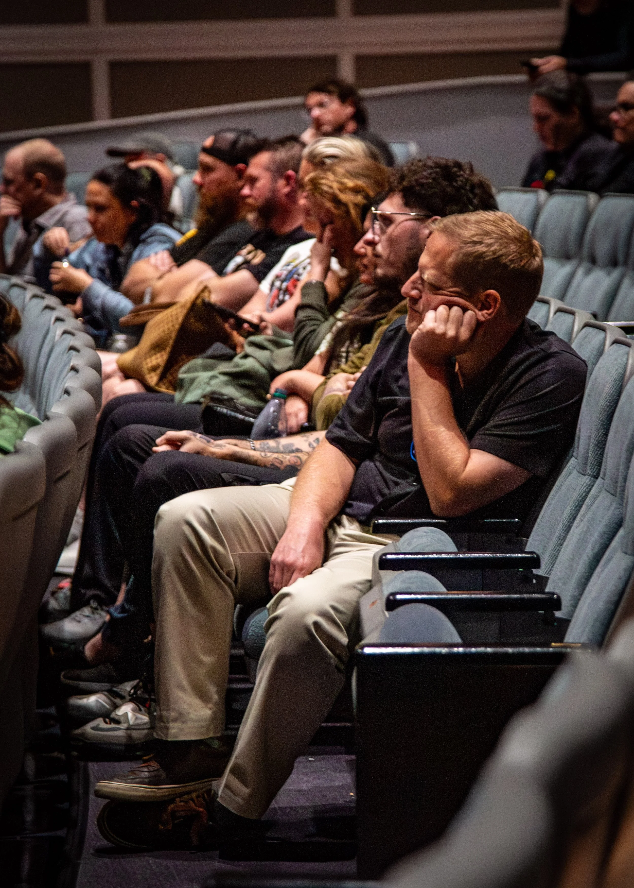 Audience sitting in theater seats, some appearing bored or distracted during a presentation or event.