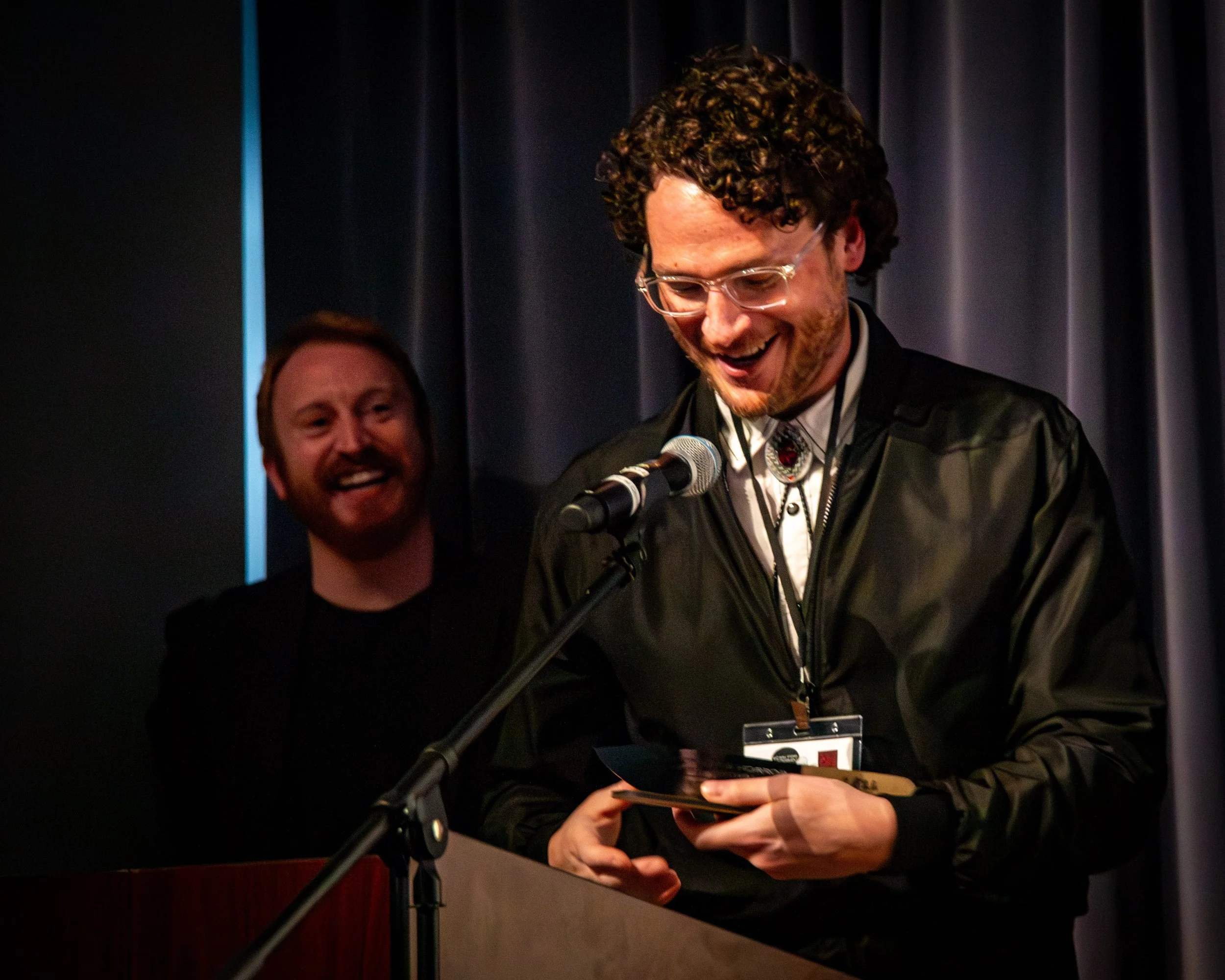 A man with curly hair and glasses is smiling while reading from a paper at a microphone, with a man in the background also smiling, in front of dark curtains.