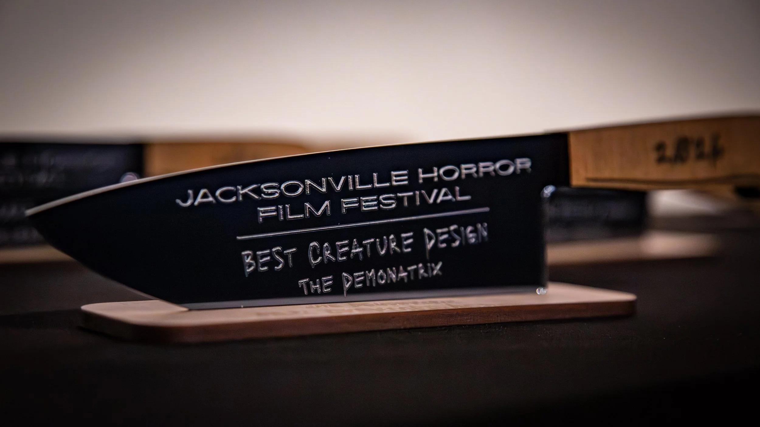Award trophy with engraved text for Jacksonville Horror Film Festival, Best Creature Design for the movie 'The Demonatrix', placed on a wooden base on a dark table.
