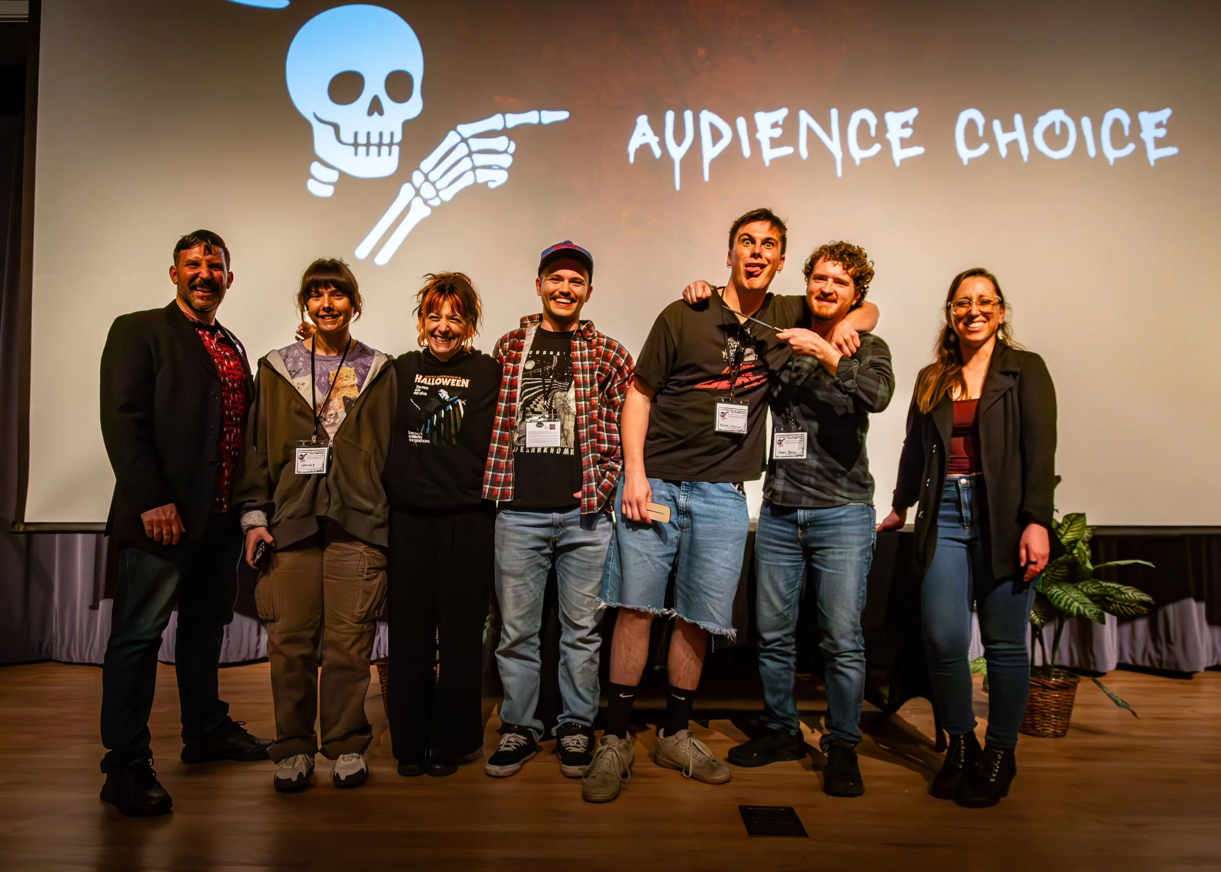 Group of seven people standing on stage in front of a large screen with a skull and pointing finger graphic and the words "Audience Choice." The individuals are smiling and embracing each other.