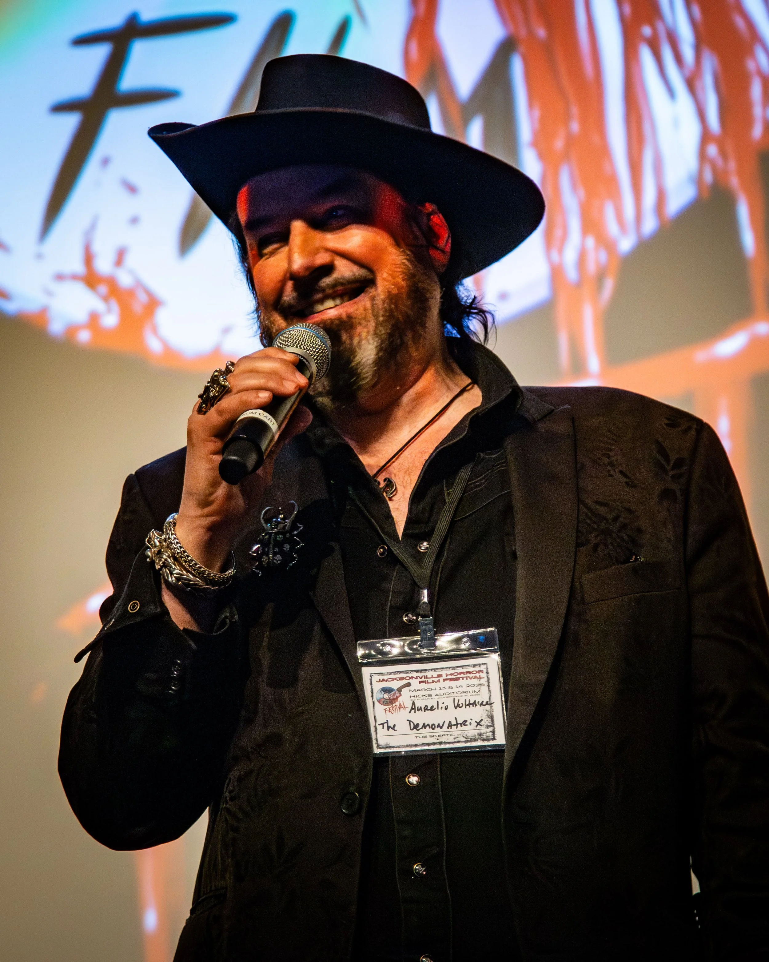 A man with a beard and long hair wearing a black hat and black jacket, holding a microphone, smiling, with a conference badge around his neck.