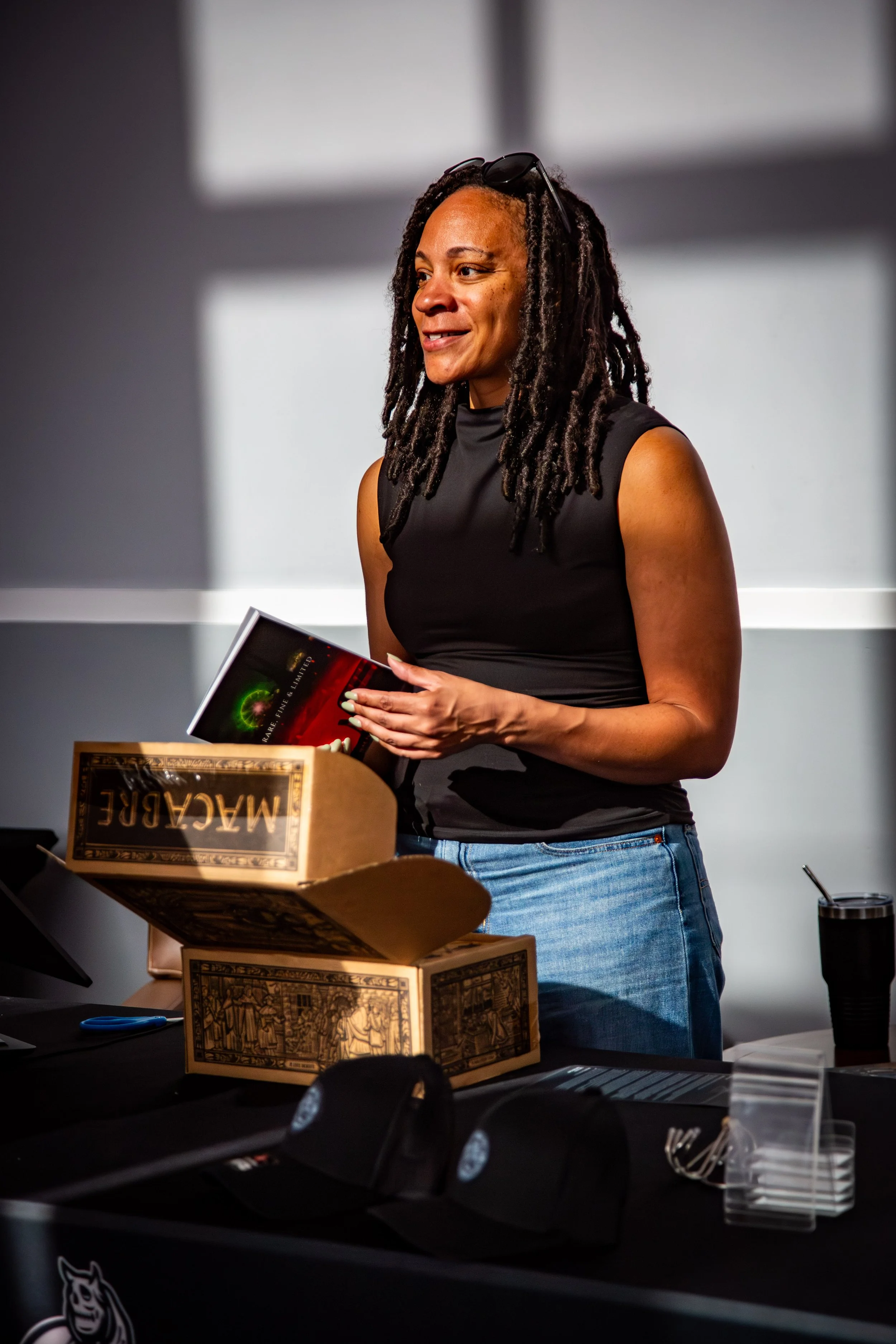 A woman with dreadlocks wearing a sleeveless black top and glasses resting on her head, smiling while holding a book, standing behind a table with a Monopoly game box, a black cup, and some plastic utensils.”}