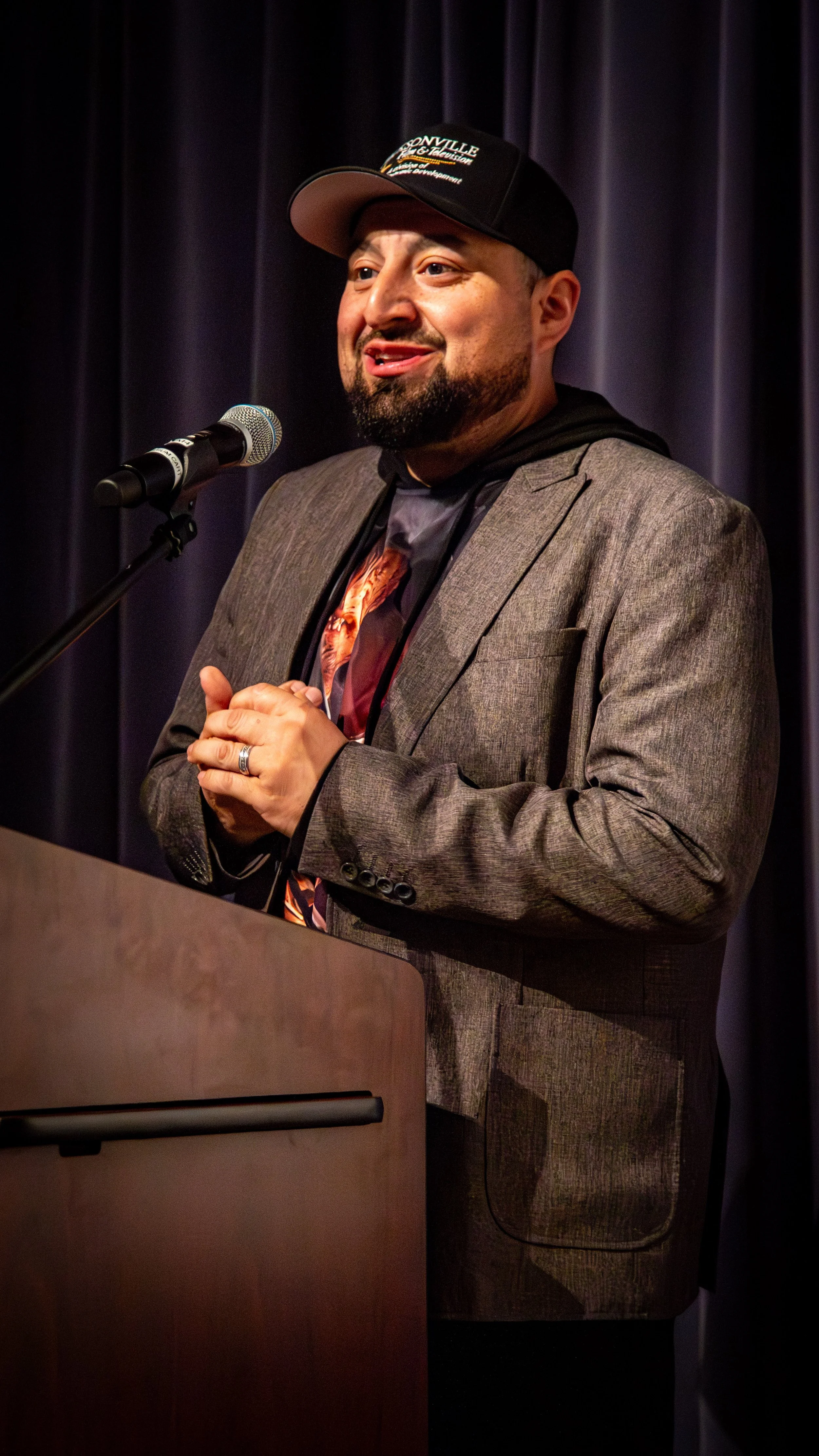 A man speaking at a podium, wearing a gray suit, graphic t-shirt, black hoodie, and baseball cap, with a microphone in front of him and a dark curtain background.