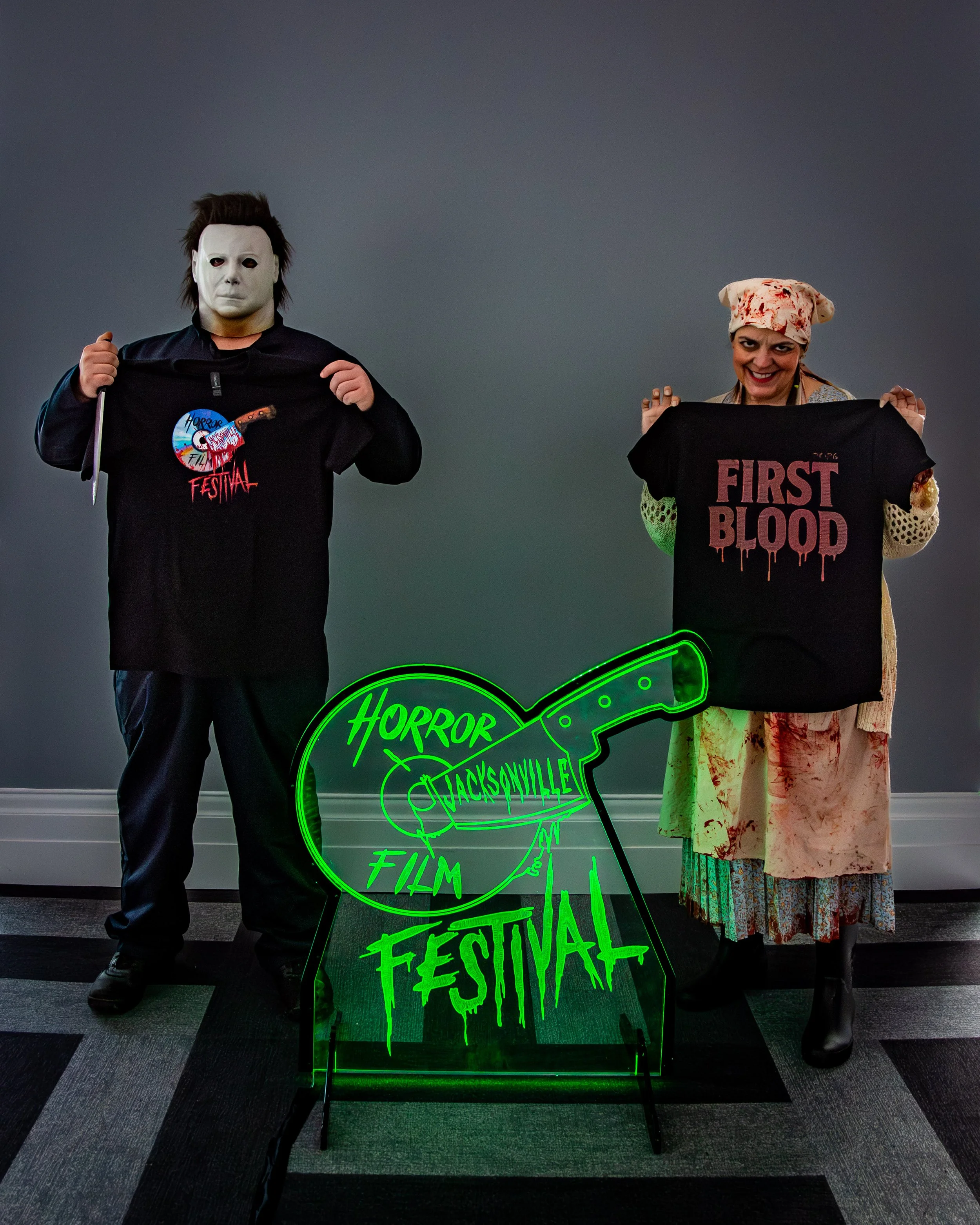 A person dressed as Michael Myers from Halloween and a woman dressed as a bloodied nurse or patient, holding horror movie T-shirts at the Horror Film Festival in Jacksonville, with a neon sign that reads Horror Film Festival.