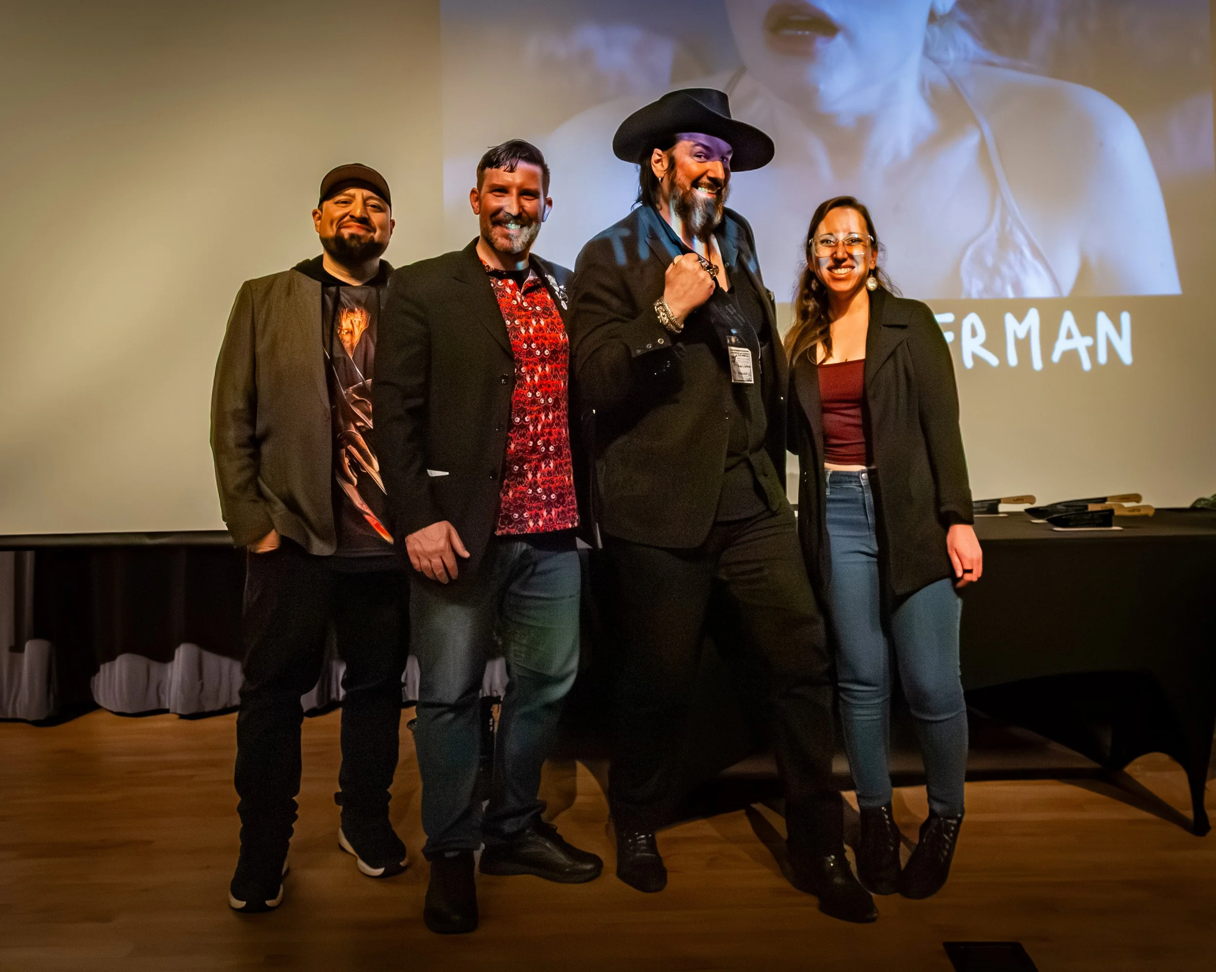 Group of four diverse people standing on stage in front of a large screen, smiling, dressed in casual and semi-formal attire, with a wooden floor and a table with some objects behind them.