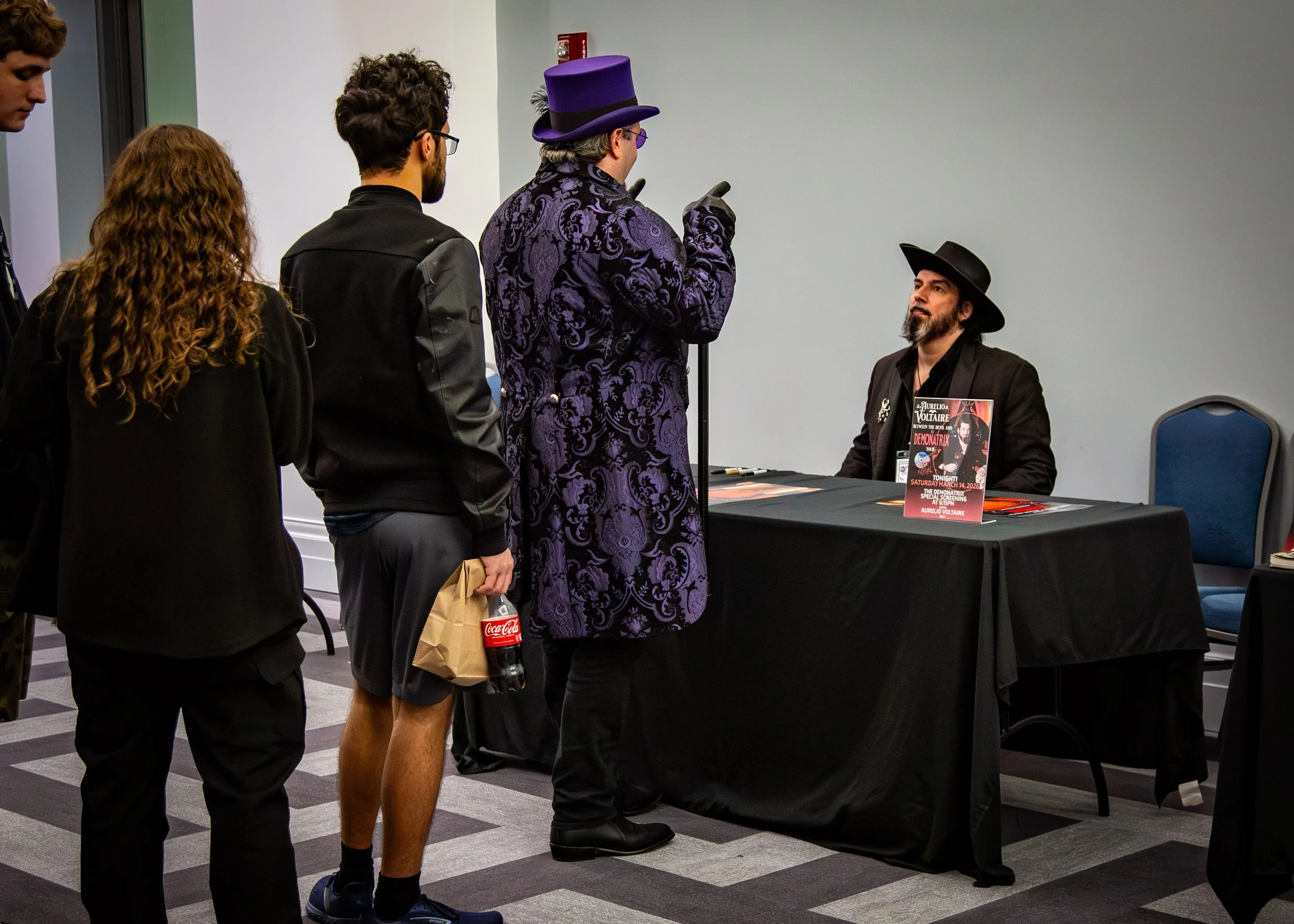 A man wearing a purple patterned coat and a matching purple top hat is standing in front of a signing table, talking to a seated man with a black hat and black clothing. Three other people are waiting in line behind him, one holding a Coca-Cola bottl